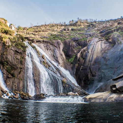 Molte cascate si riversano lungo una scogliera rocciosa in un corpo d'acqua tranquillo sotto un cielo limpido.