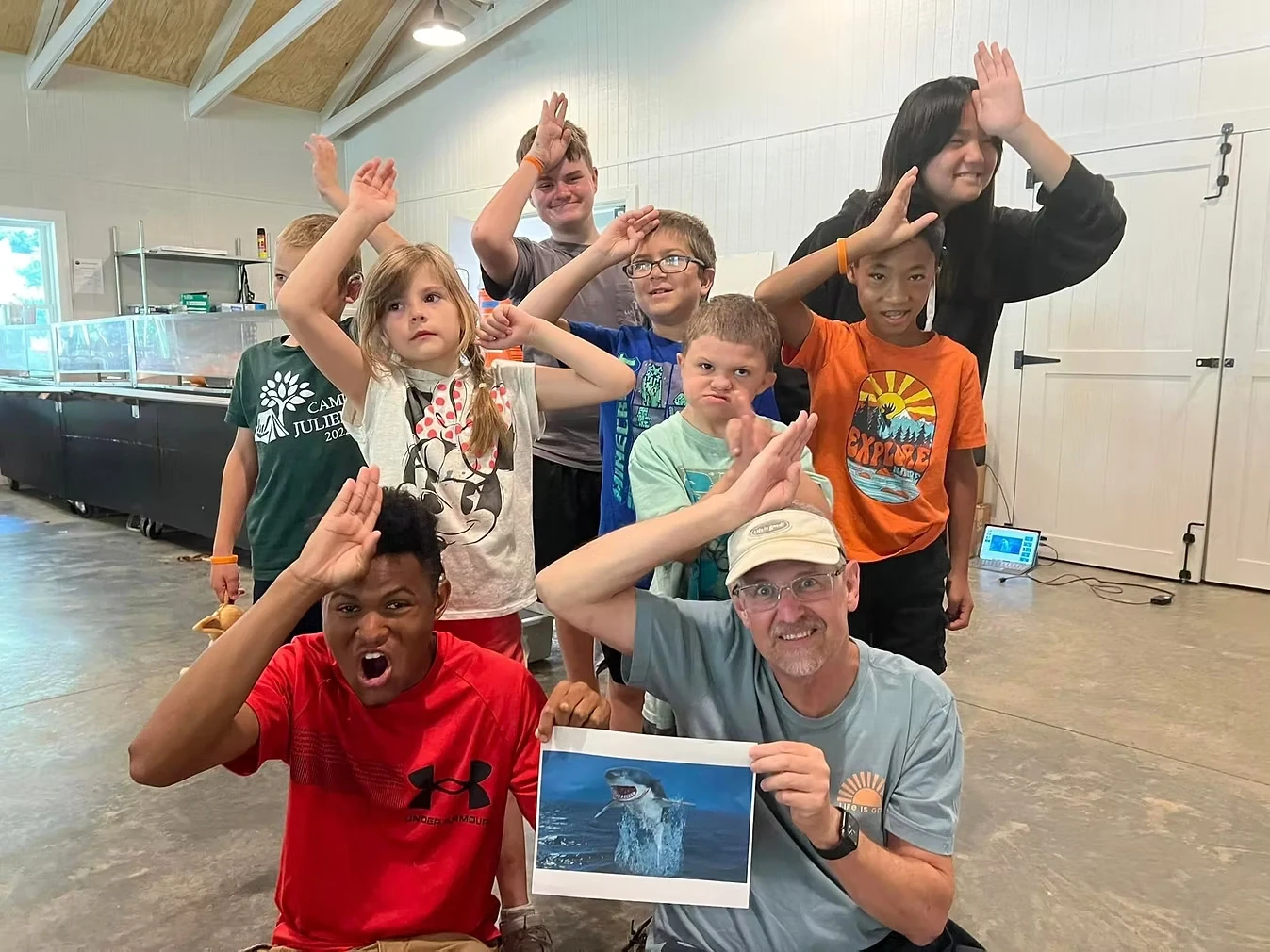 A group of campers and staff pose for a photo with a picture of a shark. They are holding their hands up to their foreheads to sign SHARK.