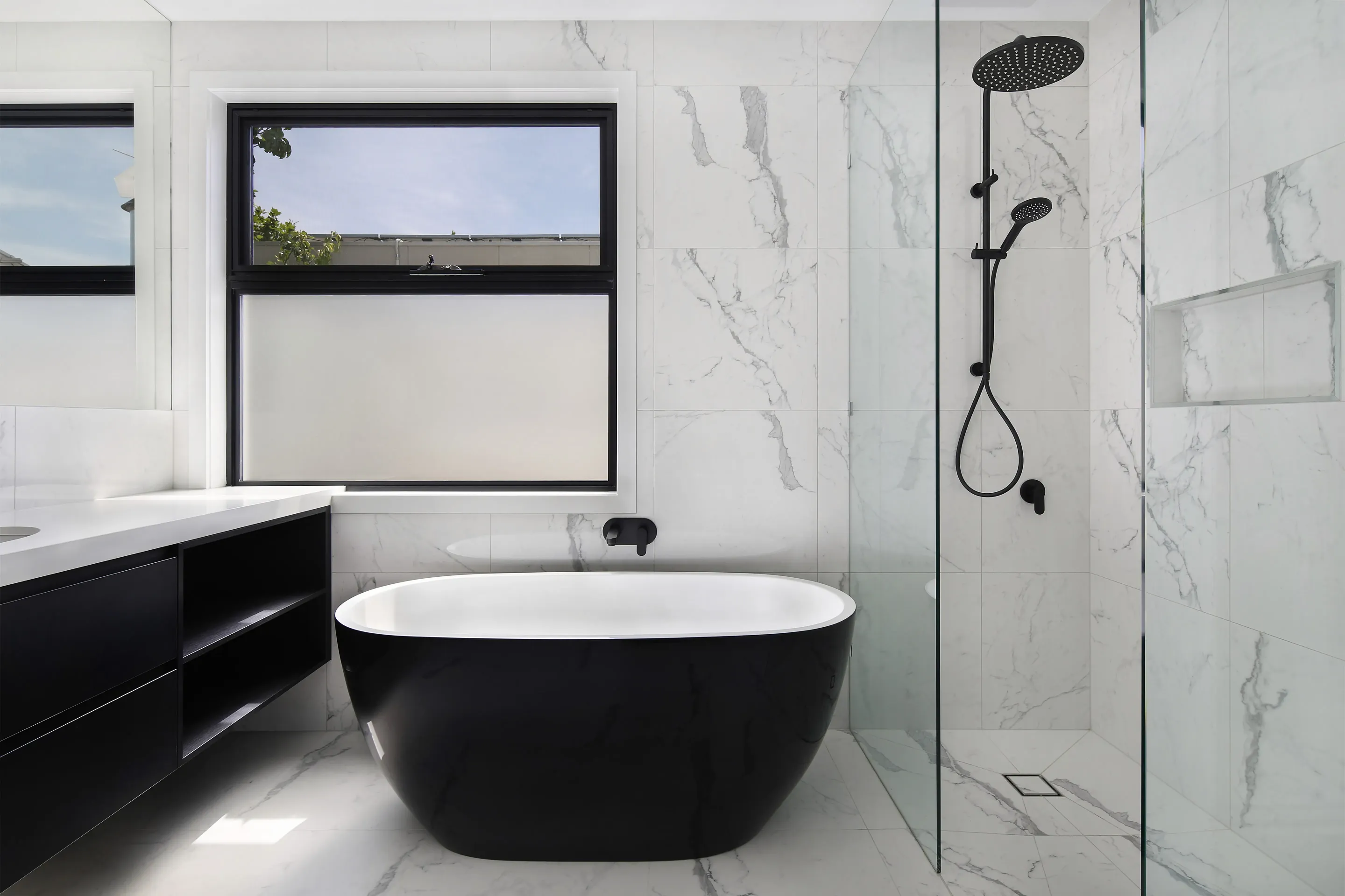 Bathroom in white marble-look tiles with a black-and-white freestanding bath, a black rail and rain shower behind a glass screen, a black floating vanity and frosted black-framed window