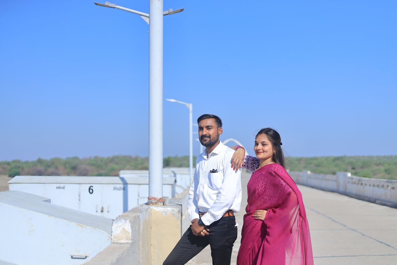 Man in a white shirt with his arm around a woman in a magenta sari beside a lamp post