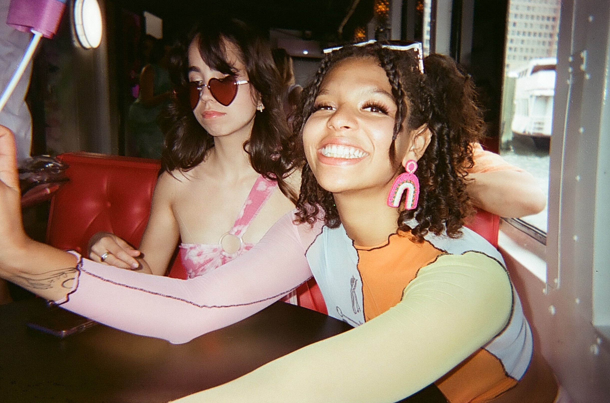 Two young women, one smiling and the other posing, sit together in a vibrant, indoor setting.