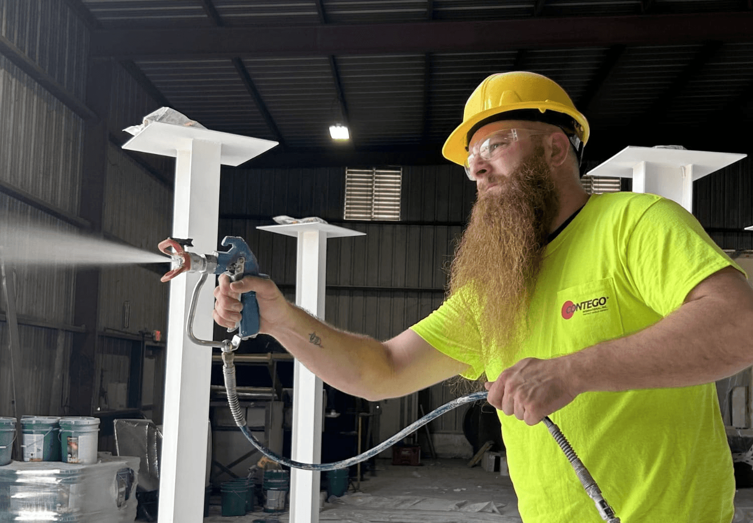 An industrial worker is applying sprayed fireproofing material onto large steel beams in a commercial building under construction, ensuring the protection of steel structures with fire resistive materials to maintain their structural integrity. The process involves using a spray application technique that enhances passive fire protection against high temperatures.