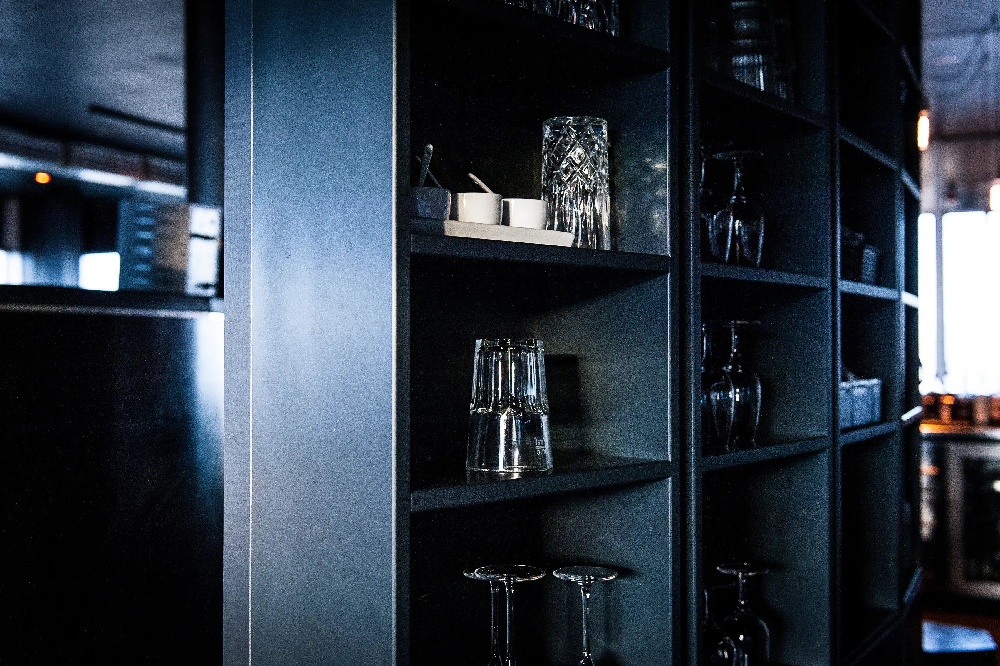 Dark bar shelving at SOLAR SKY LOUNGE with crystal and plain glasses, small ceramic bowls and upside-down wine glasses in a black metal rack.