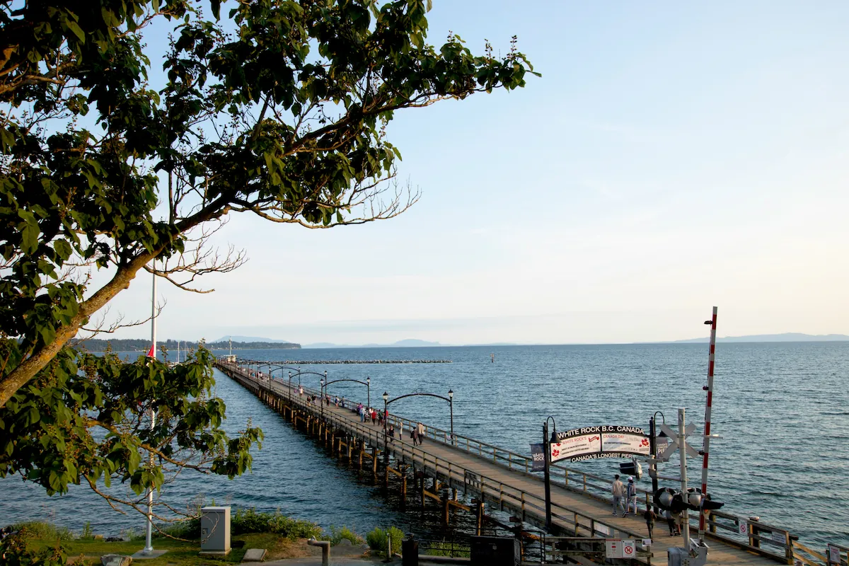 White Rock Pier extending over the ocean at sunset, with visitors walking along the boardwalk and shoreline views of White Rock, BC.