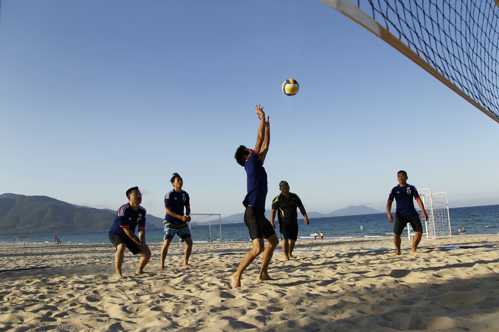 A beach volleyball player in blue sets the ball while teammates watch on a sandy court near the ocean.