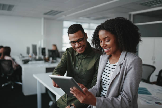 Two African business professionals in an office discussing information on a tablet, highlighting teamwork, digital collaboration, and modern workplace culture.