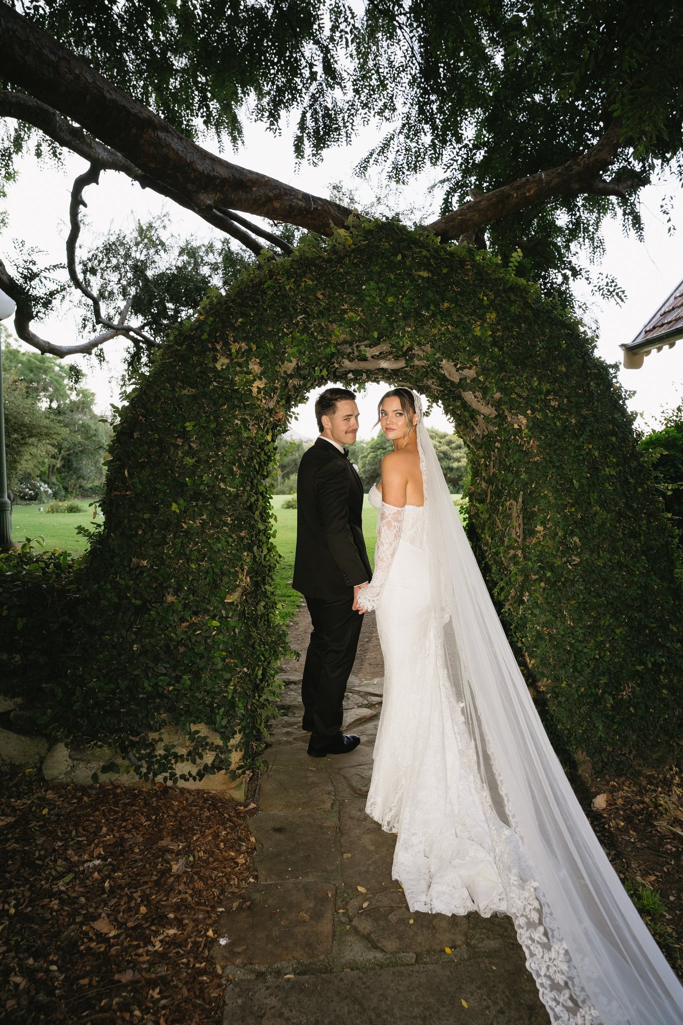 Bride and groom holding hands under an ivy-covered garden archway with overhanging tree branches