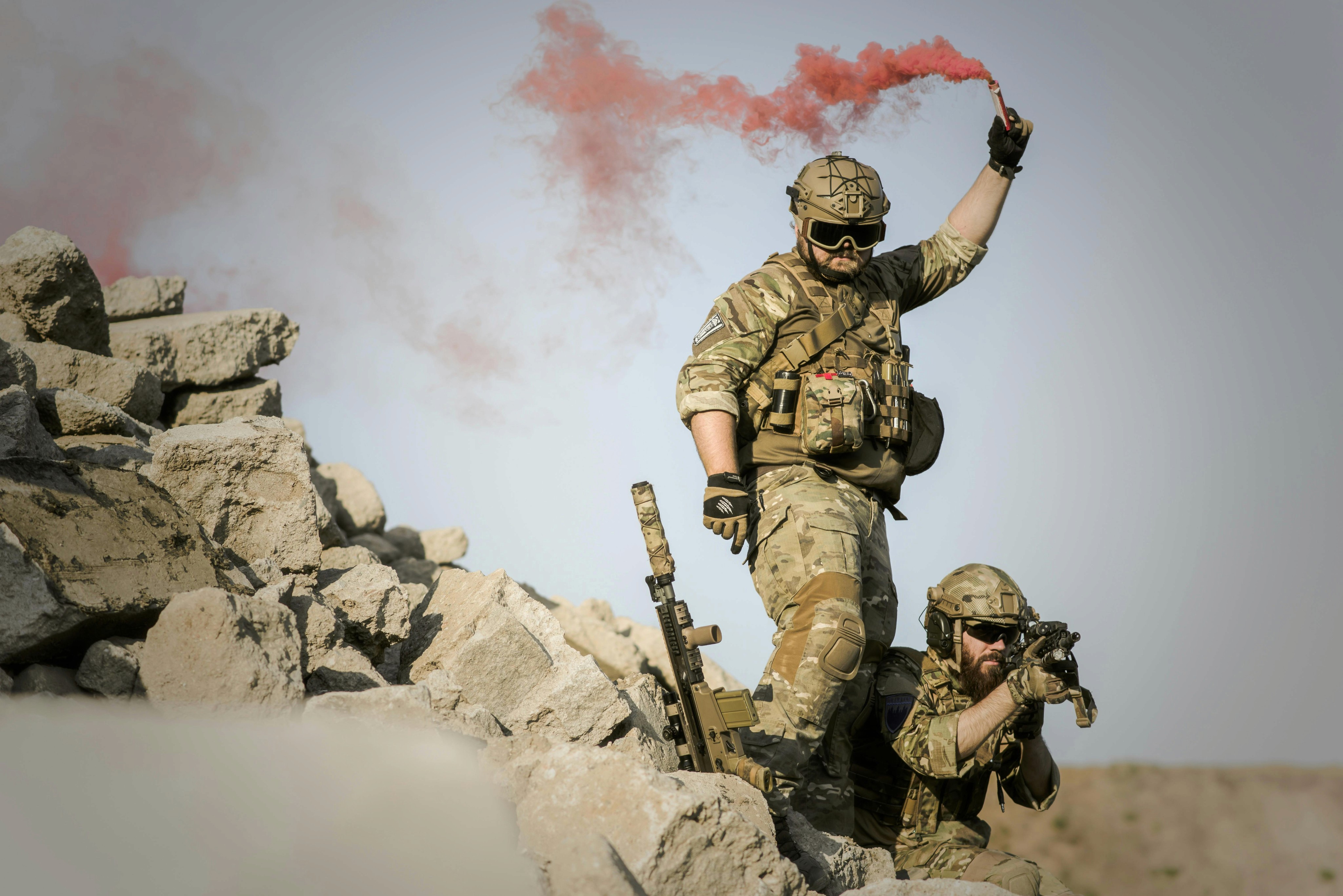 Two tactical operators in desert/tan camouflage uniforms positioned among large rocky debris. The standing soldier is holding a red smoke flare high above his head, creating dramatic red smoke trails against the light sky. Both personnel are wearing tactical helmets with protective eyewear and full combat gear including knee pads and gloves. The second operator is partially visible in a lower position, holding a tactical rifle. The setting appears to be a desert or rubble-filled training environment.