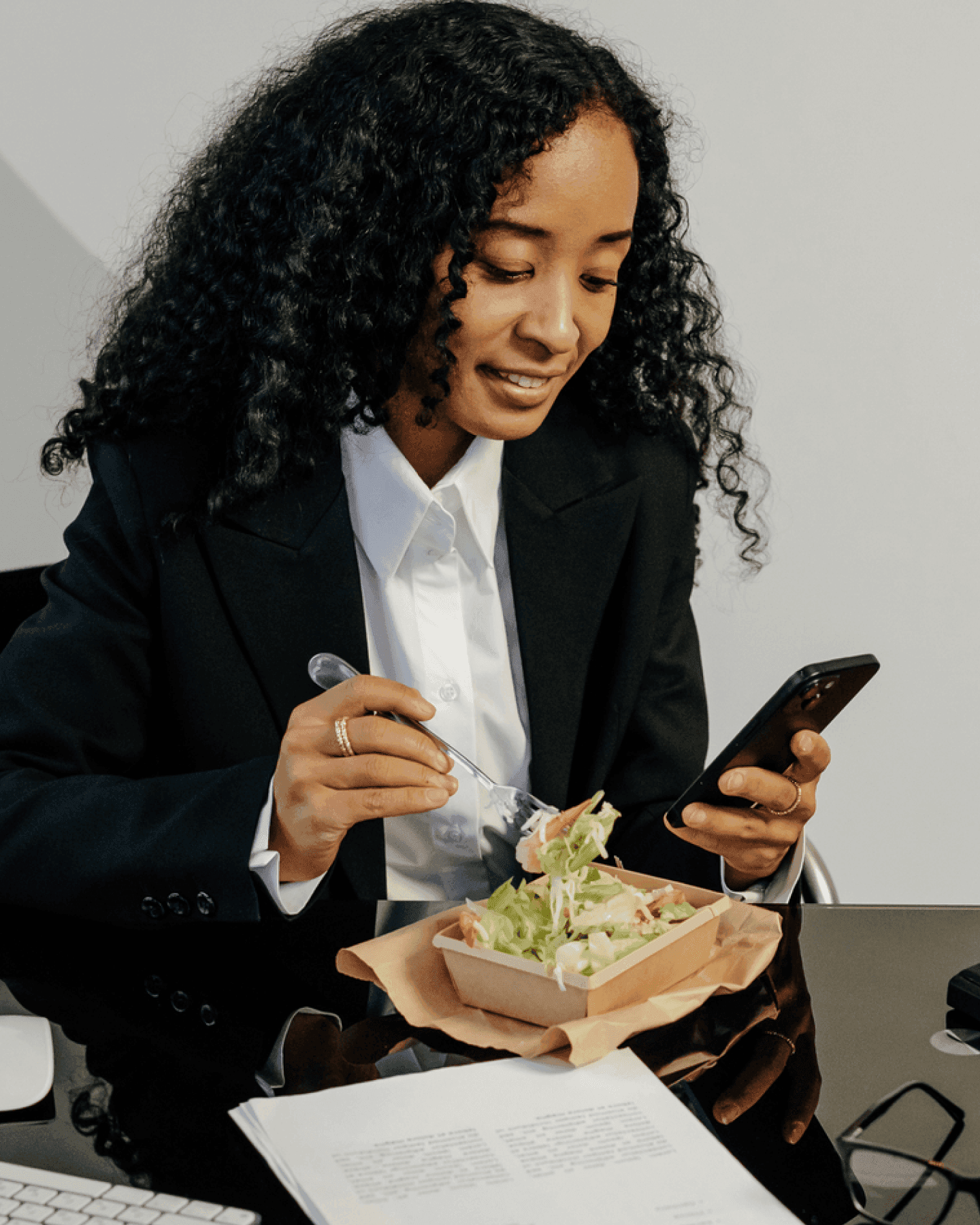 Woman in a suit eating a salad while using a smartphone at a sleek black desk.