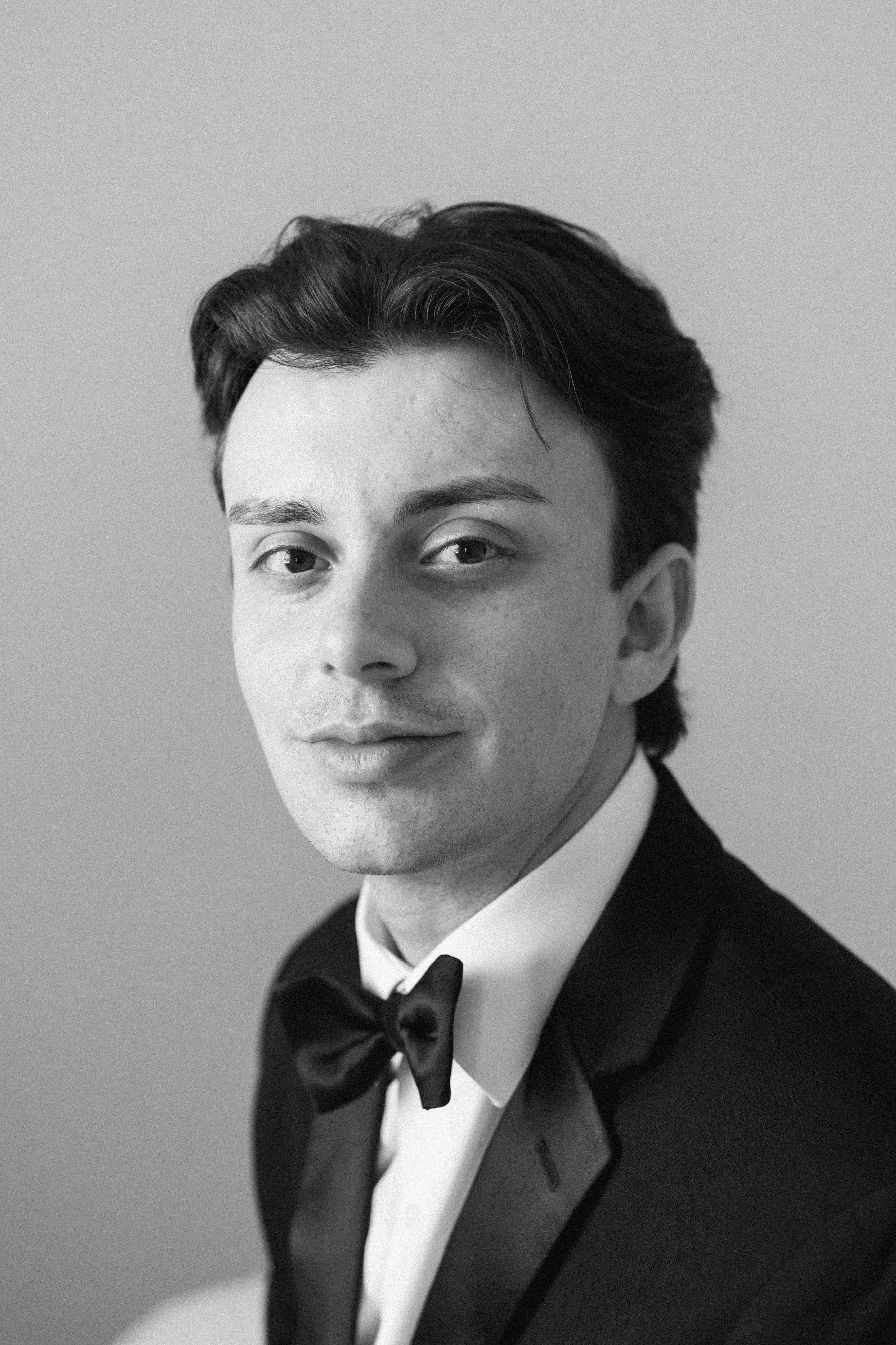 A black-and-white portrait of a man in a tuxedo and bow tie, looking directly at the camera.