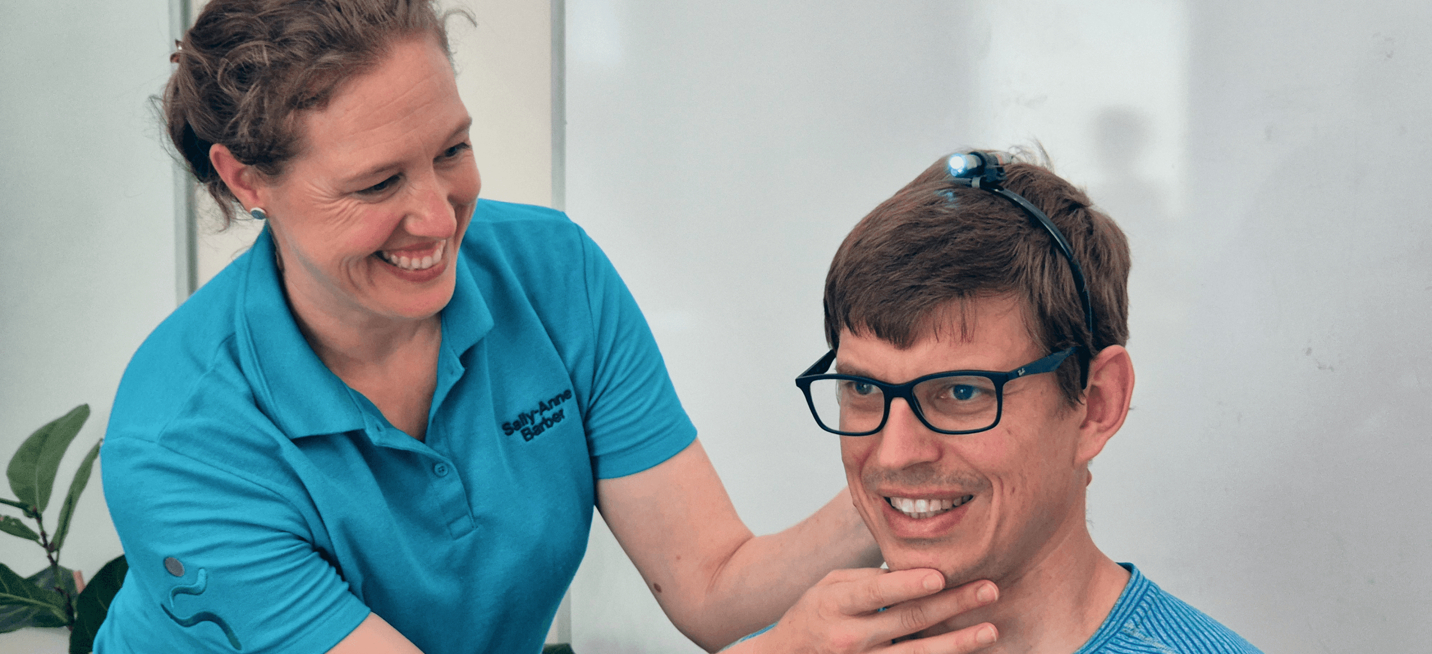 A smiling woman adjusts the chin of a young man wearing glasses, both dressed in blue shirts.