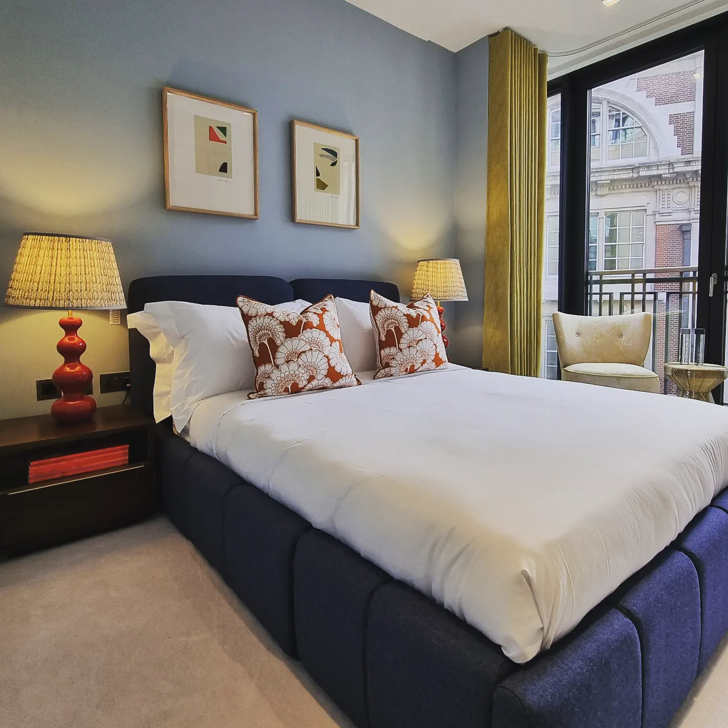 A modern hotel room features a neatly made bed with white linens and patterned pillows, flanked by stylish red bedside lamps on wooden nightstands, set against a backdrop of contemporary artwork and large windows with mustard yellow curtains.