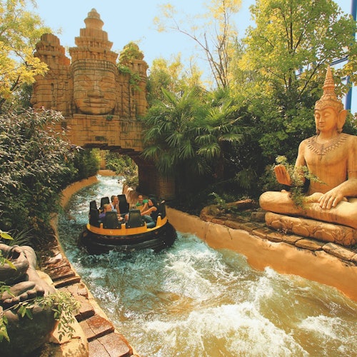 אנשים ברפסודה משייטים דרך נחל מים עם נושא טרופי, עוברים ליד גילופי אבן של פנים ופסל גדול של בודהה ישוב.