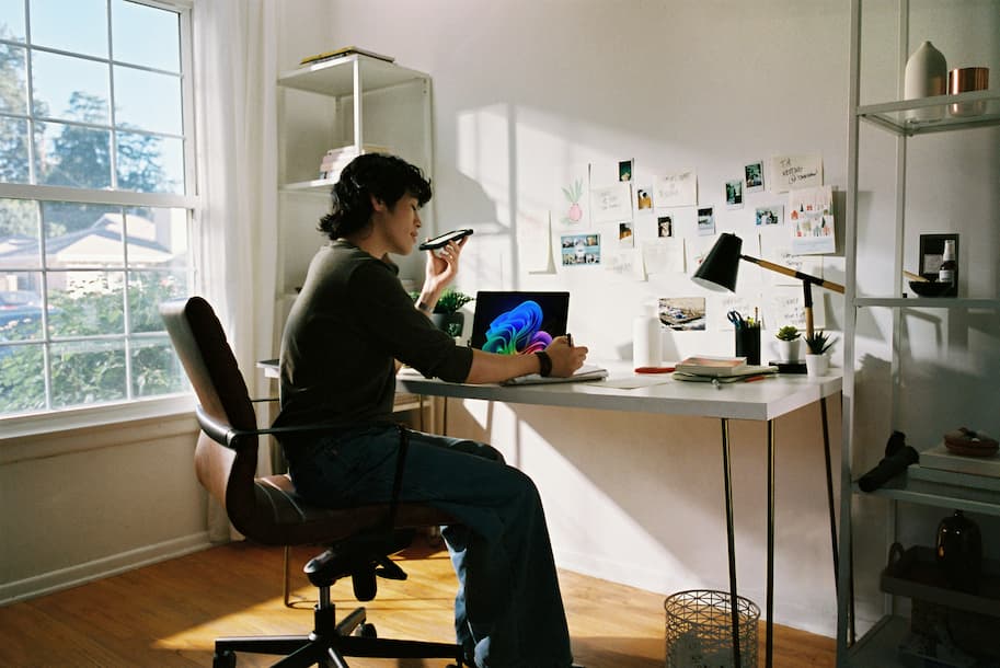 A professional working at a white desk in a sunlit office, speaking on a phone while managing tasks on a laptop.
