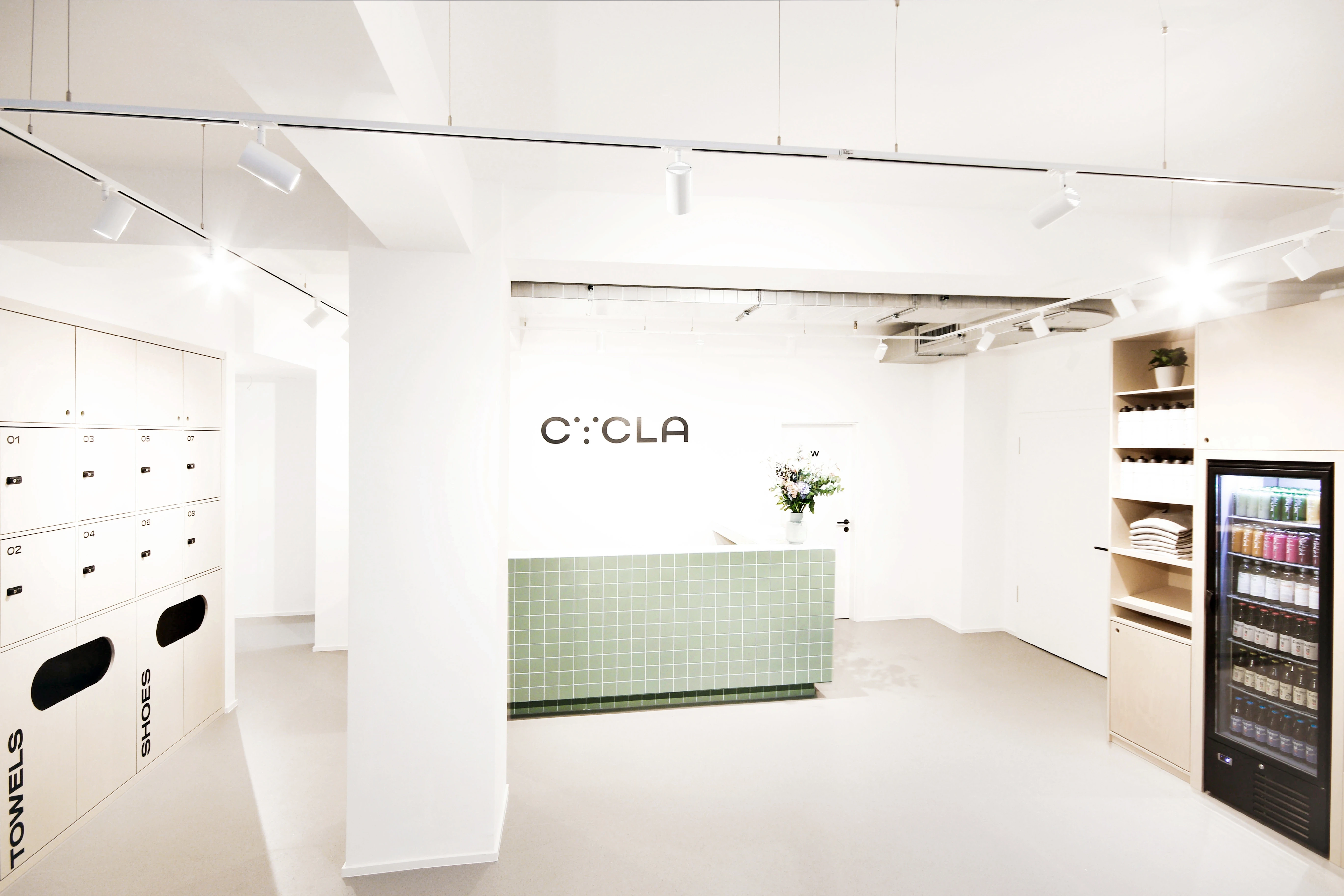 Entrance area of the CYCLA studio in white with a mint-green tiled reception counter, black CYCLA logo on the wall, towel and shoe lockers and a drinks fridge in a wooden shelf.