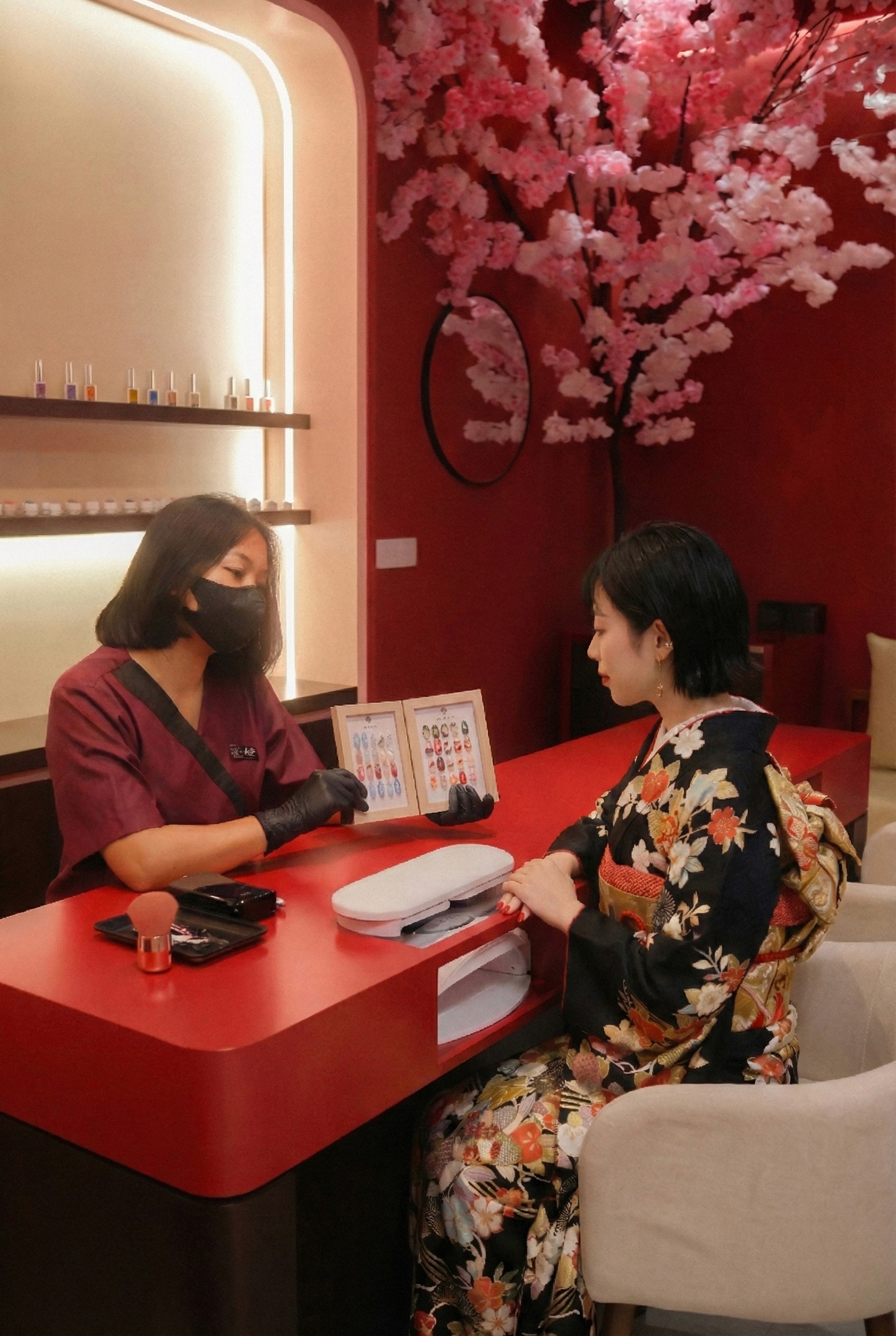Woman in kimono getting nails done, selecting polish with technician at nail salon. Cherry blossom decor and red interior.