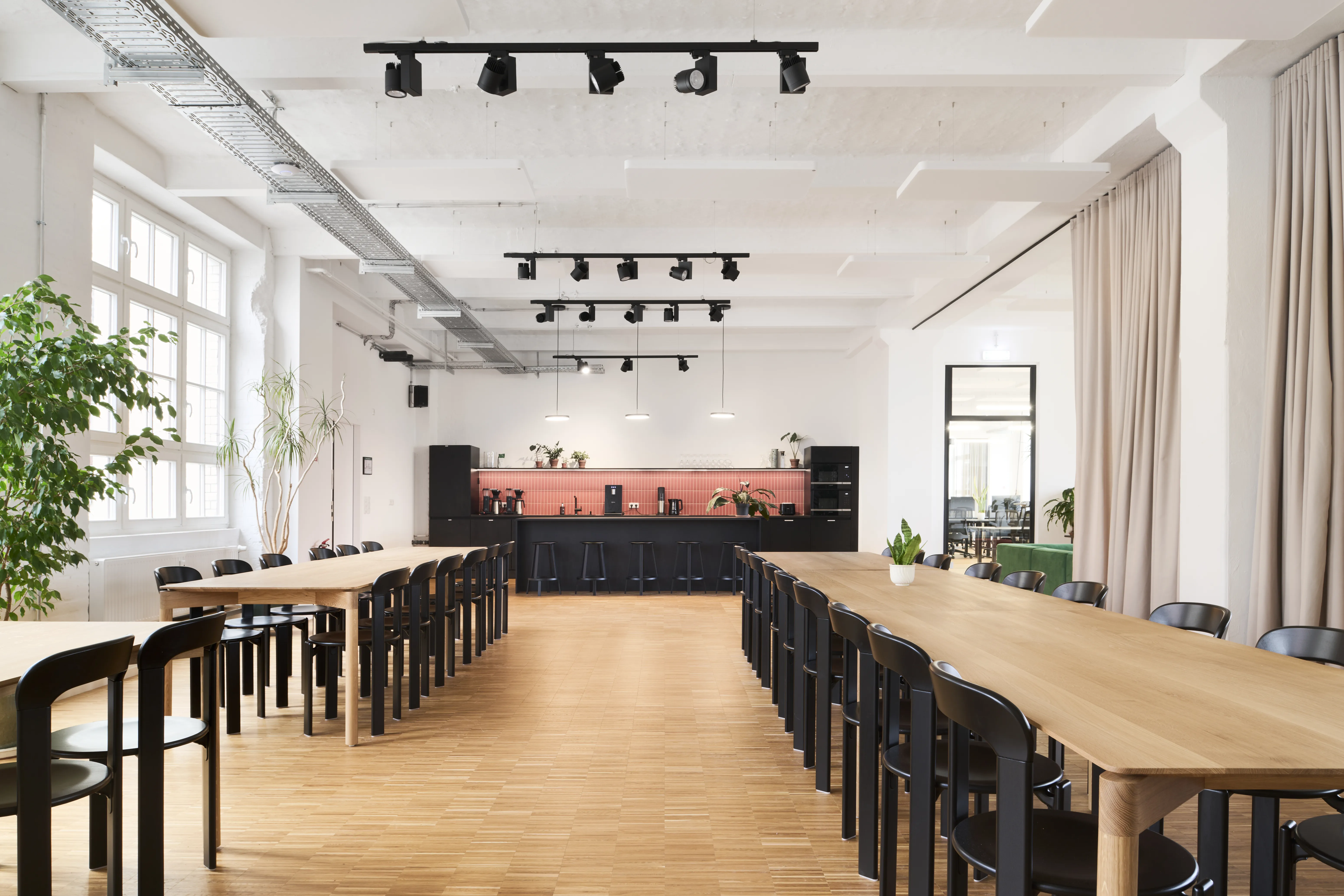 Canteen at PLAN A with two long oak tables and black chairs, a black kitchen wall with coral-red tiled splashback, beige curtains and rows of black spotlights under a white beamed ceiling.
