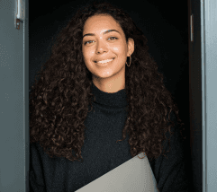 A smiling woman with curly hair stands in a doorway, holding a laptop or binder. She wears a black sweater.