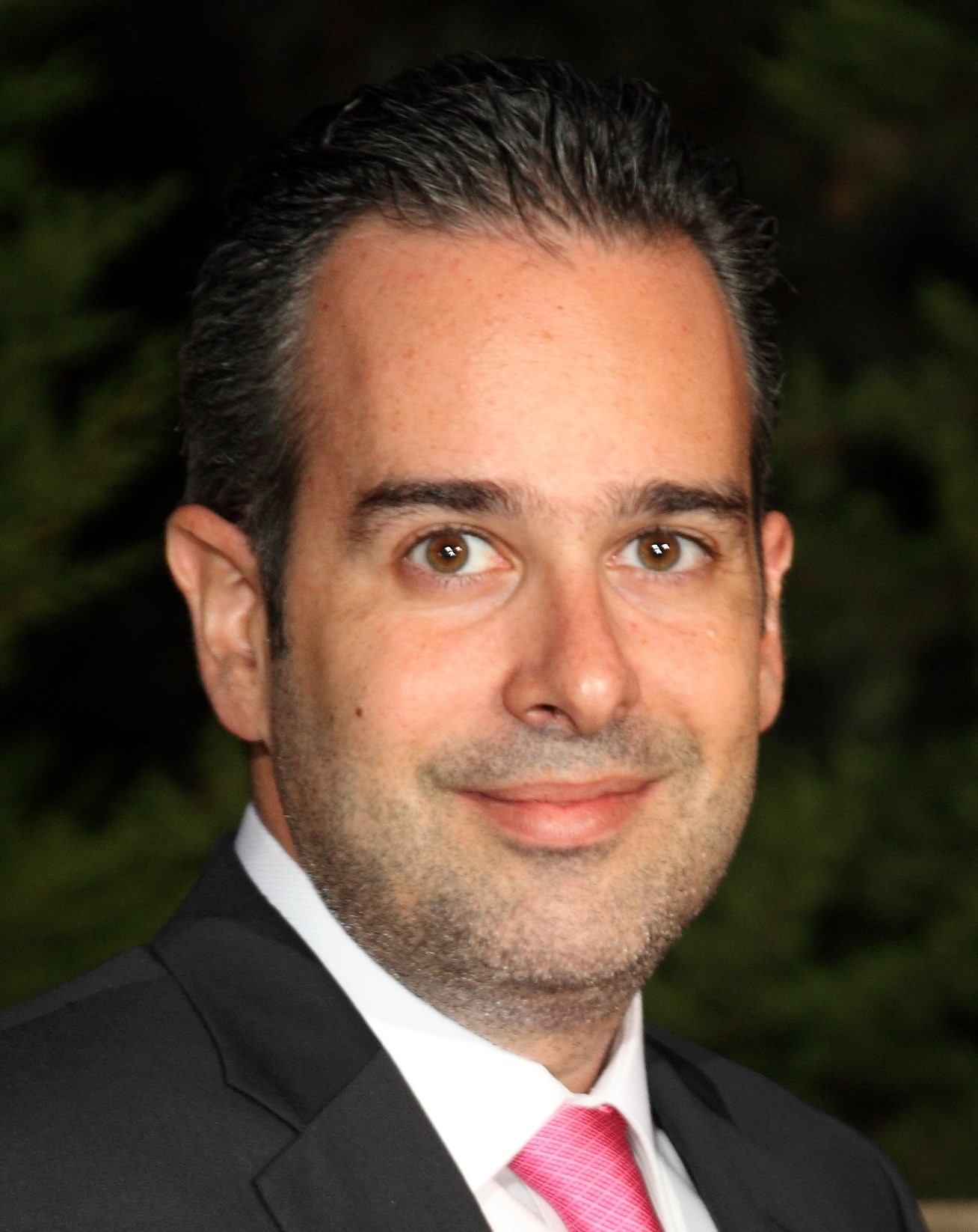 A smiling person wearing glasses and a patterned shirt stands against a plain white background.