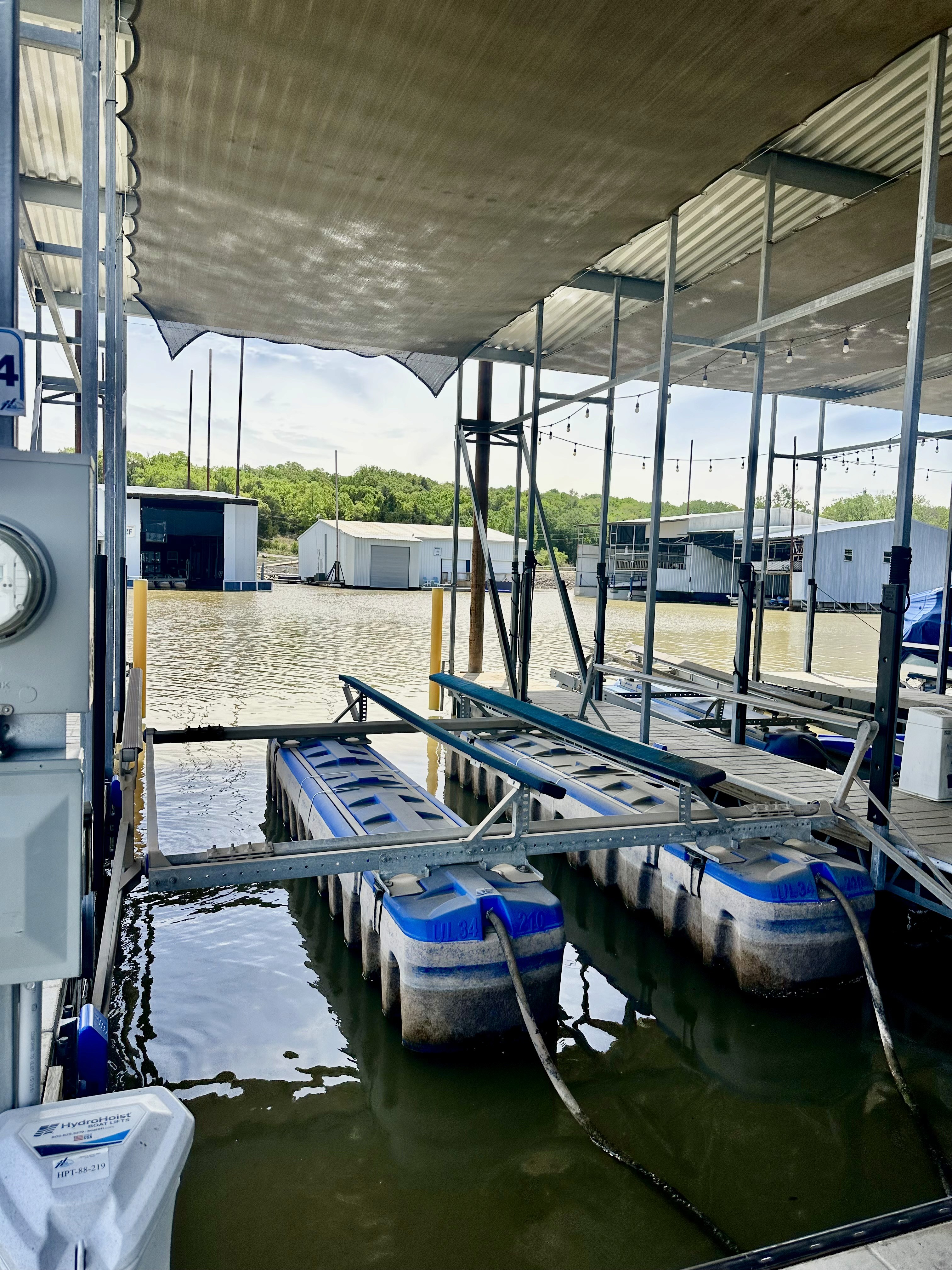 The image depicts a floating dock with a metal boat lift system situated on a calm, scenic lake, under a metal canopy and surrounded by lush greenery in the background.