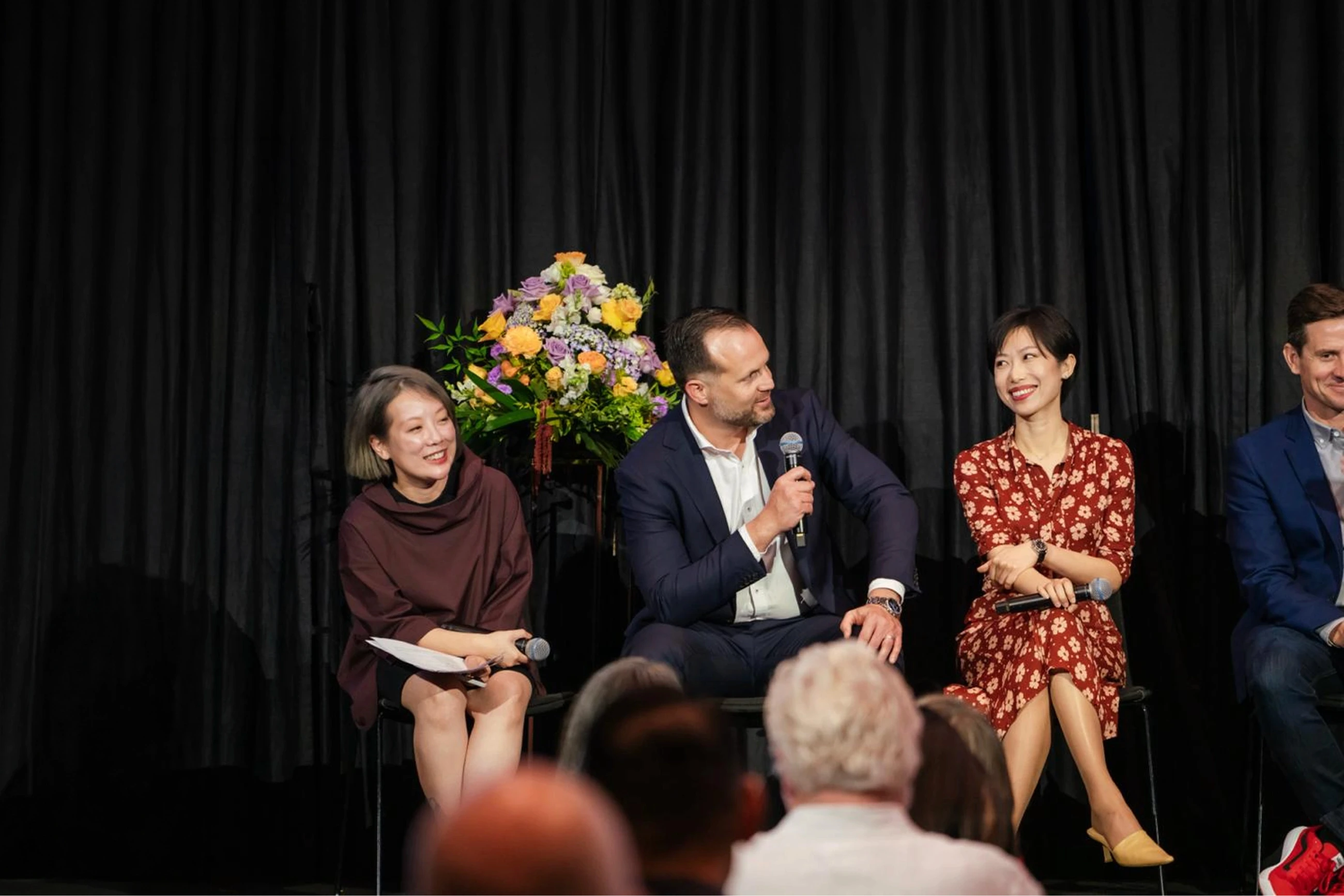 A panel of three individuals sits on stage, with two women and one man, dressed in formal attire, engaging in conversation, in front of a dark curtain backdrop and a vibrant floral arrangement on a stand.