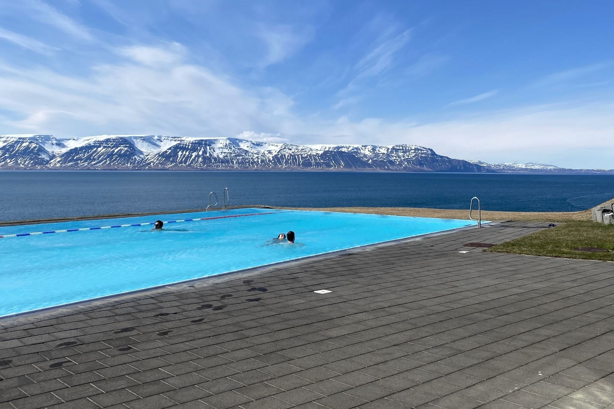 An aquamarine swimming pool on the edge of a paved area with the sea beyond, and snow-capped mountains on the other side of the bay. Iceland.