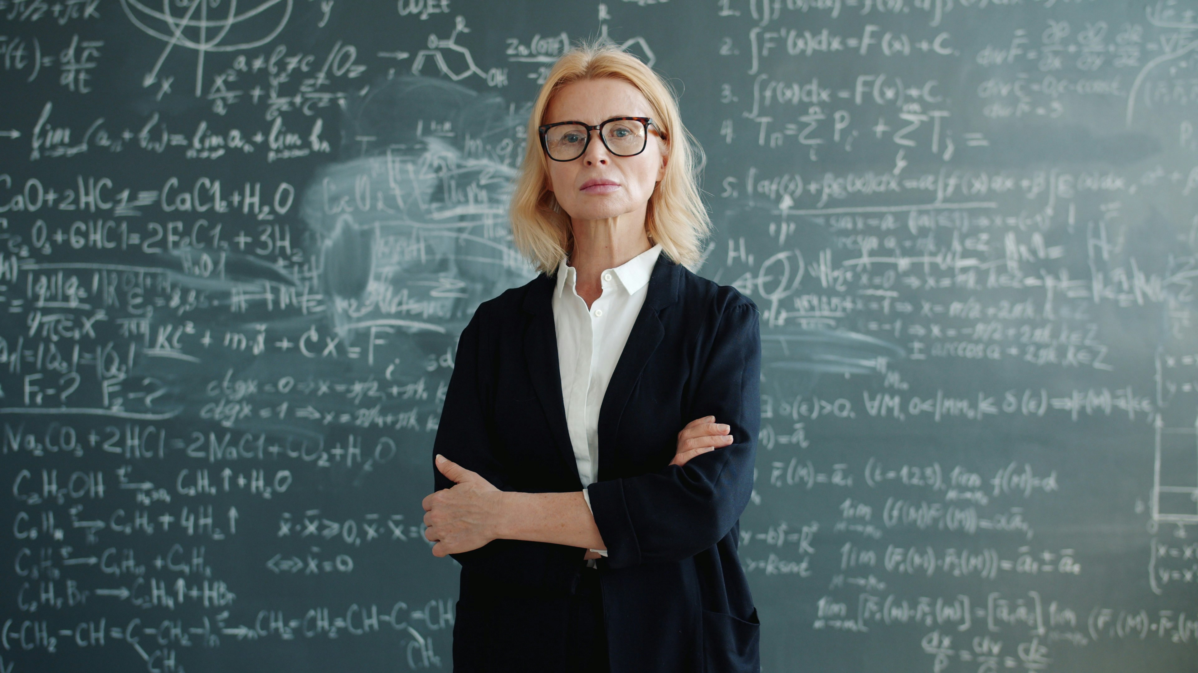 Woman in glasses stands before a chalkboard with equations.