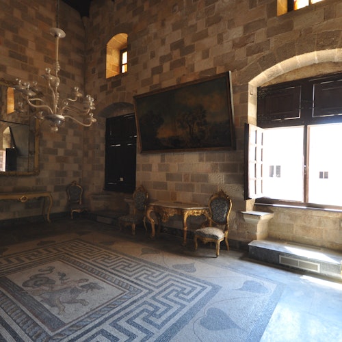 A historic room with stone walls, ornate chairs, a chandelier, a large painting, and a mosaic floor lit by sunlight from tall windows.