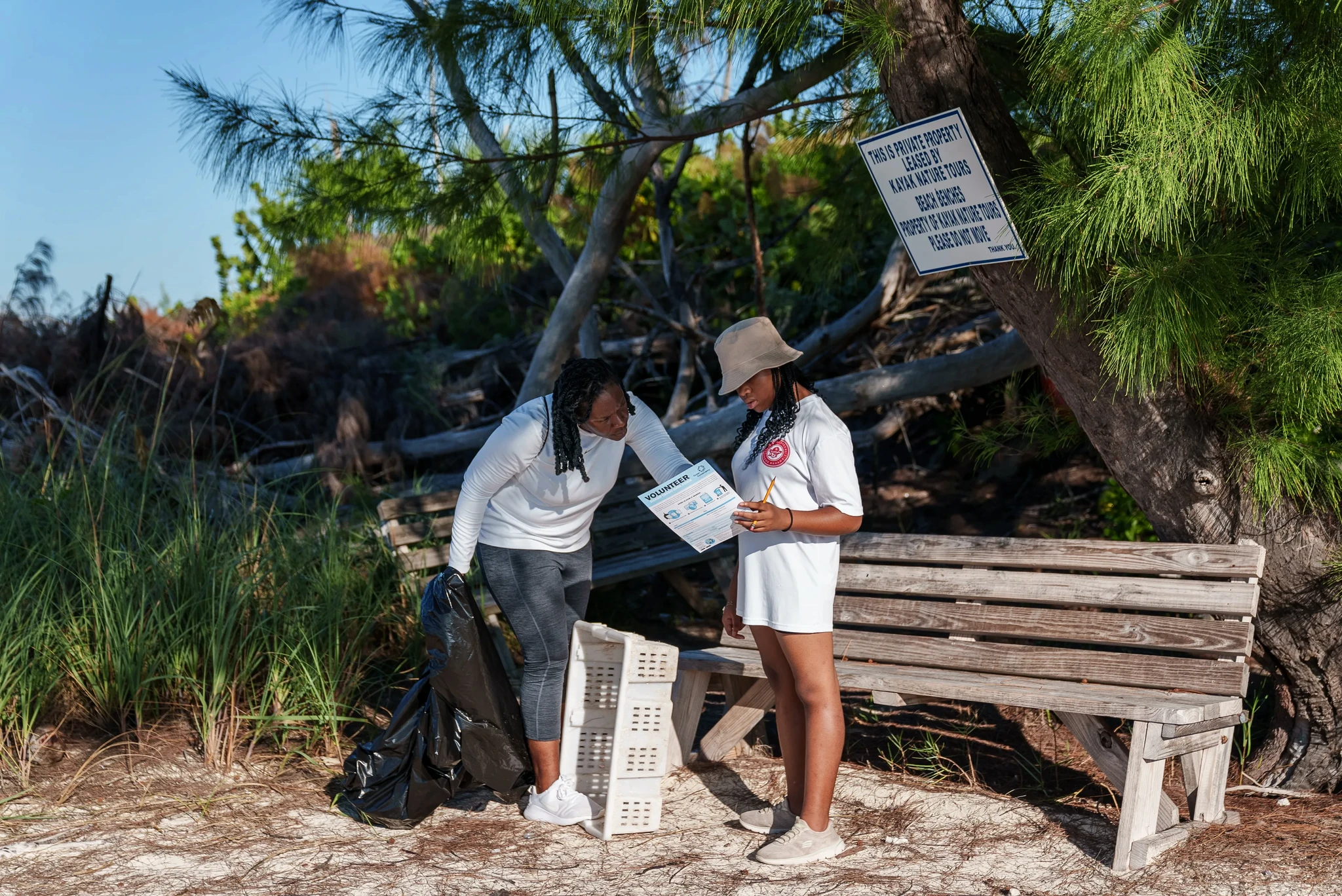 Two people standing near a beach bench, reviewing a document while participating in a coastal cleanup activity with a trash bag and crate nearby.