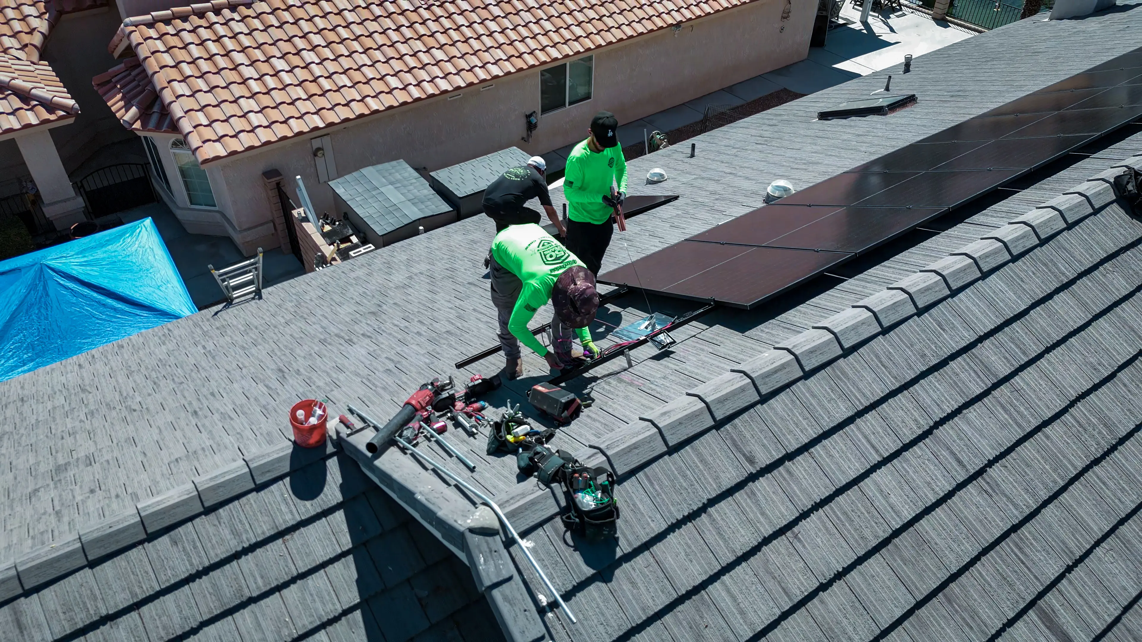 Two installers kneeling on a shingle roof, laying out panels and power tools along the ridge