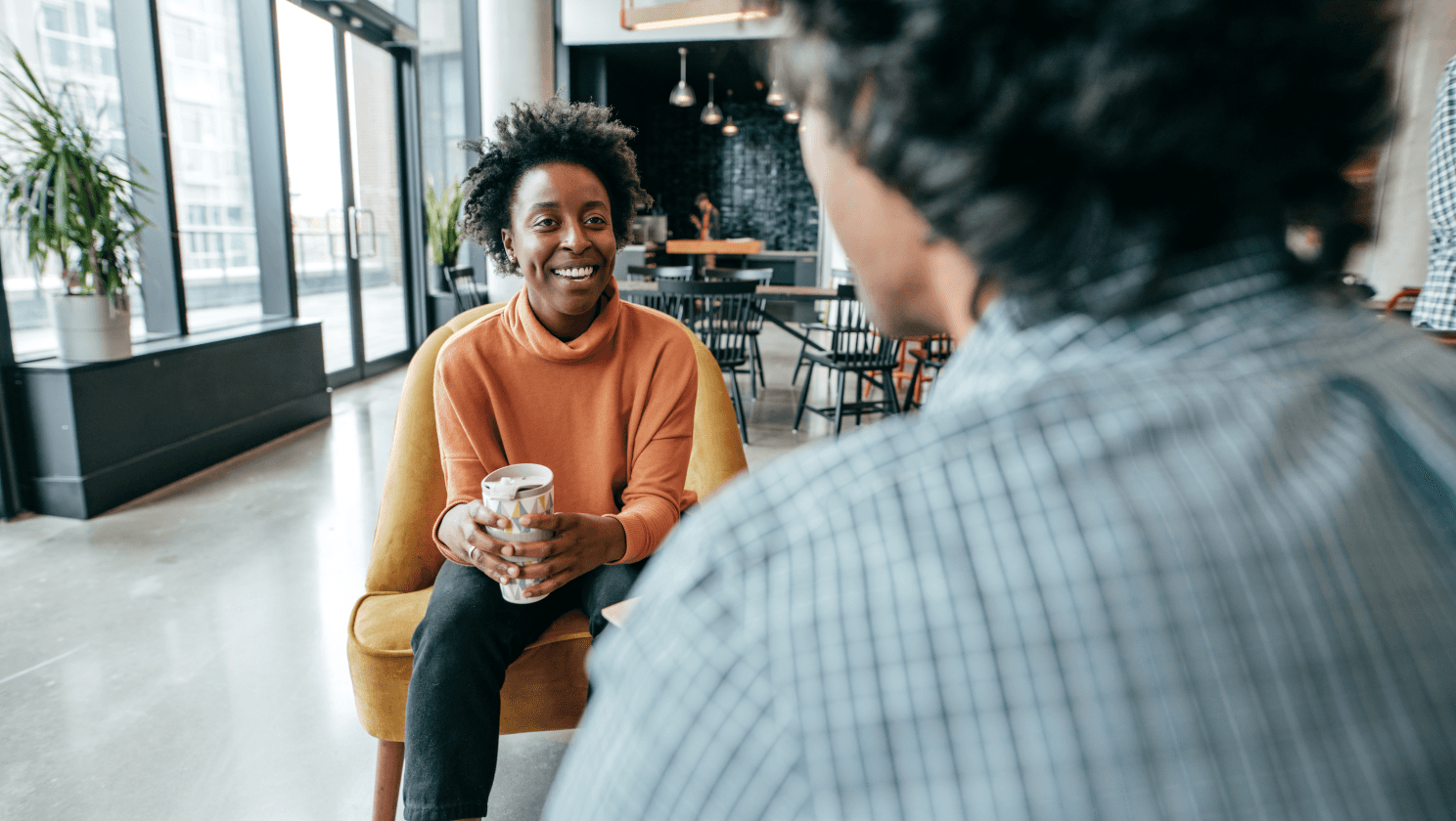 Person holding a coffee cup in orange turtleneck smiling at person sitting in front wearing a plaid blue green shirt 