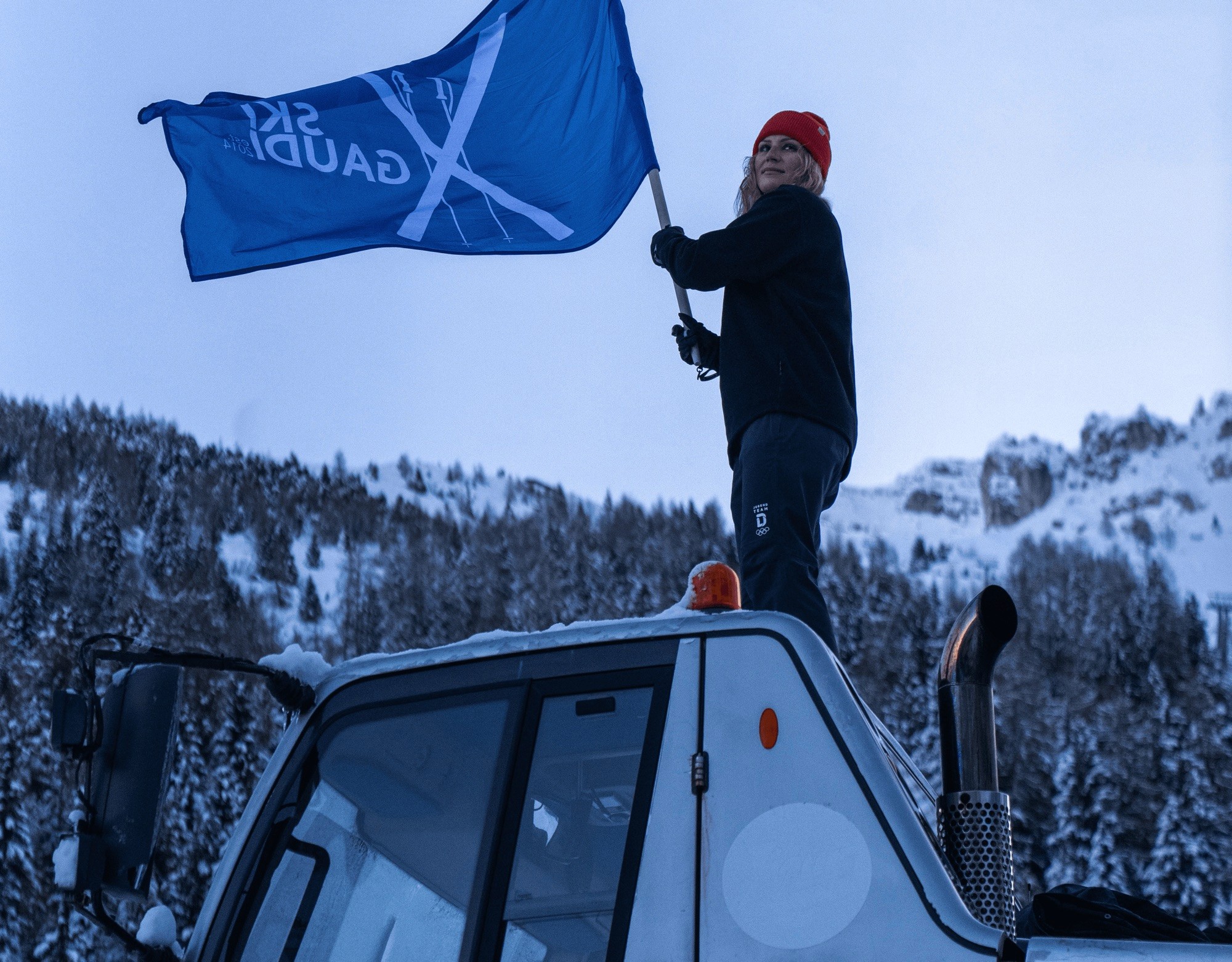 Person holding blue flag on snow groomer in snowy mountain landscape.
