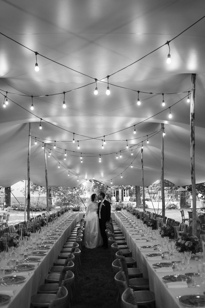Sposi camminano sotto le stringhe di luci nel lungo tavolo del ricevimento di matrimonio, bianco e nero, reportage Fotolize