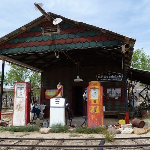 老旧加油站配有三台复古汽油泵，一座带有 B.F. Goodrich 招牌的木制建筑，前方则是铁轨。