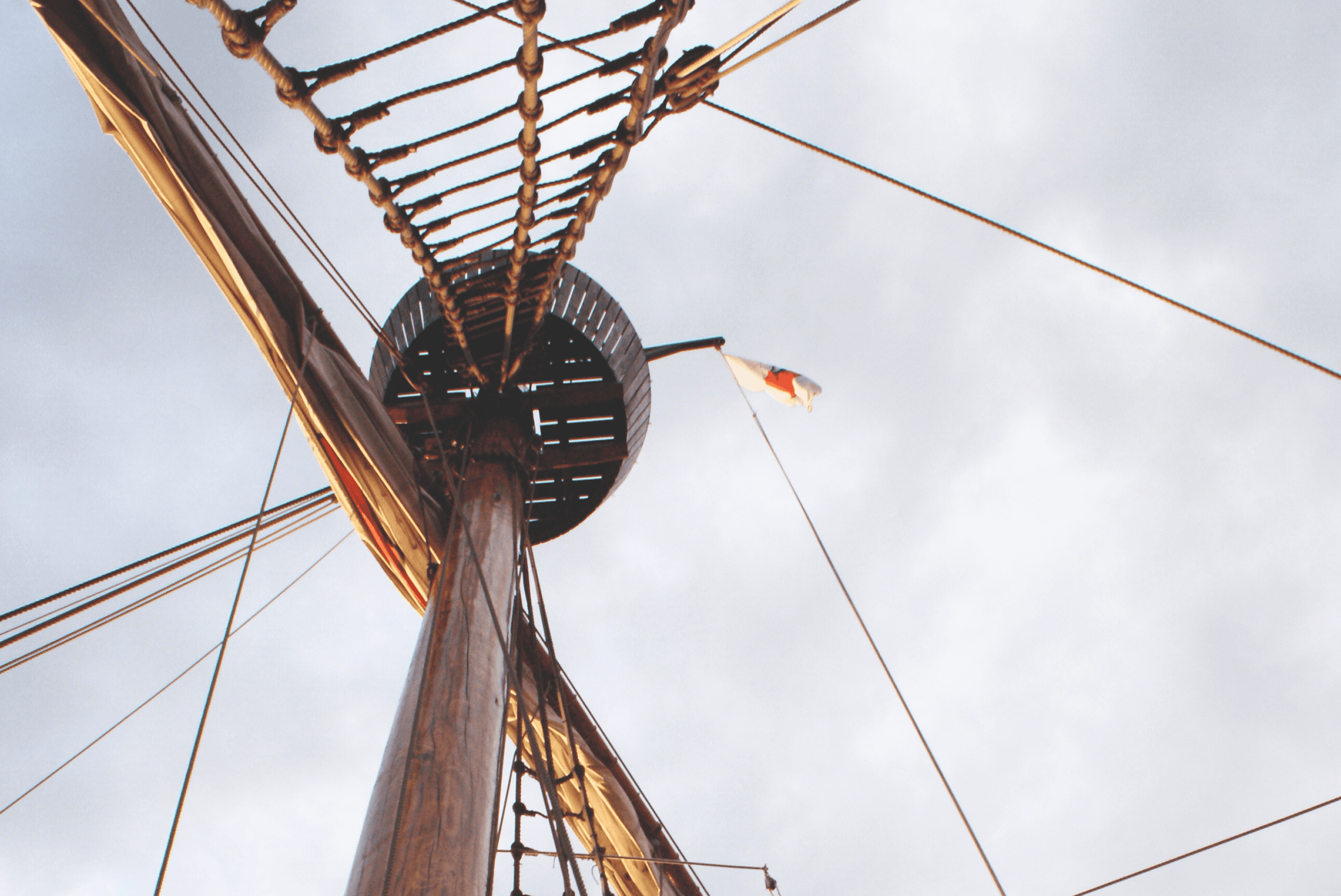 Ship mast with rigging and flag against sky.