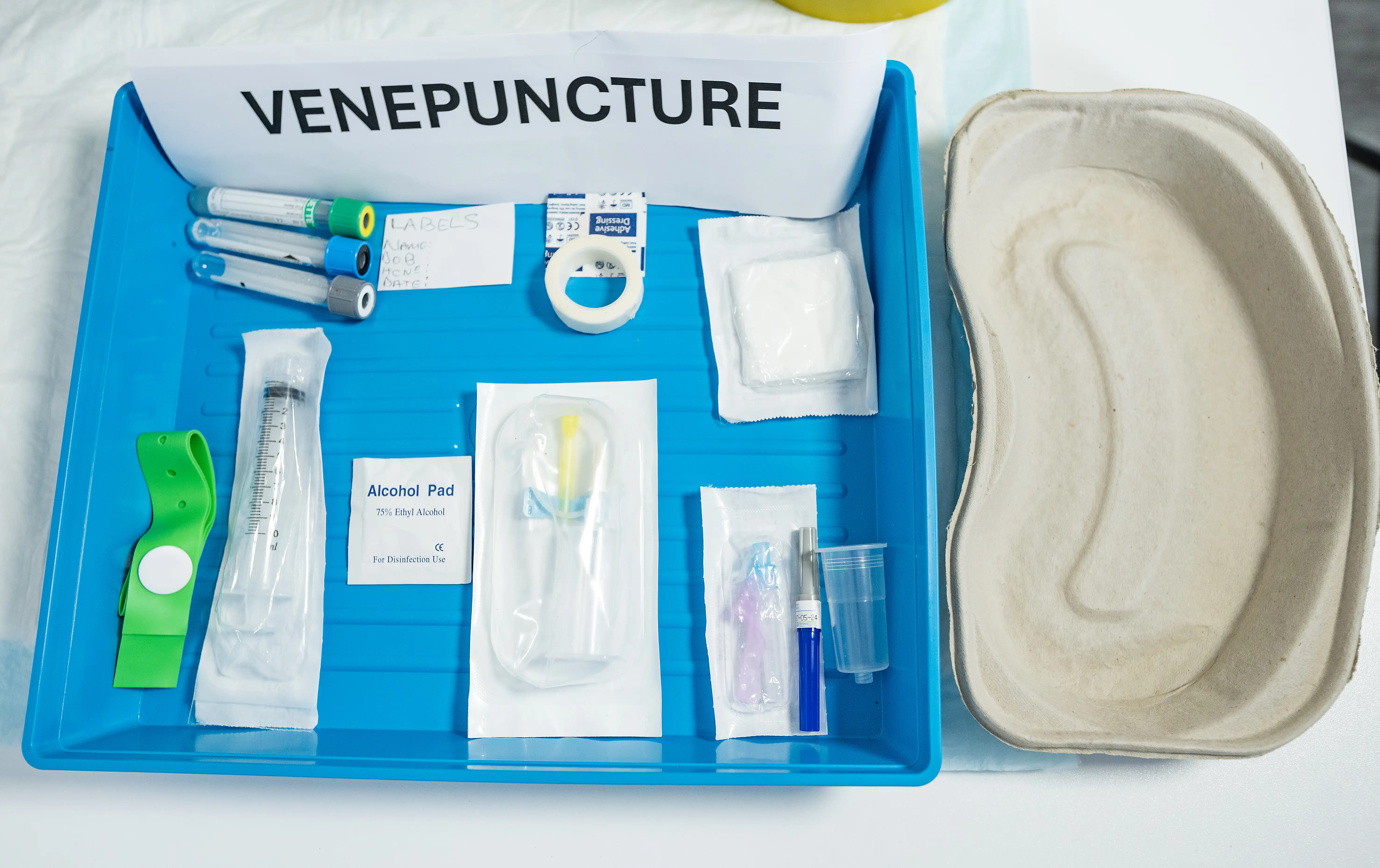 Tray containing venepuncture equipment including blood collection tubes, alcohol pad, tourniquet, syringe, and sterile medical supplies, laid out for a clinical procedure.