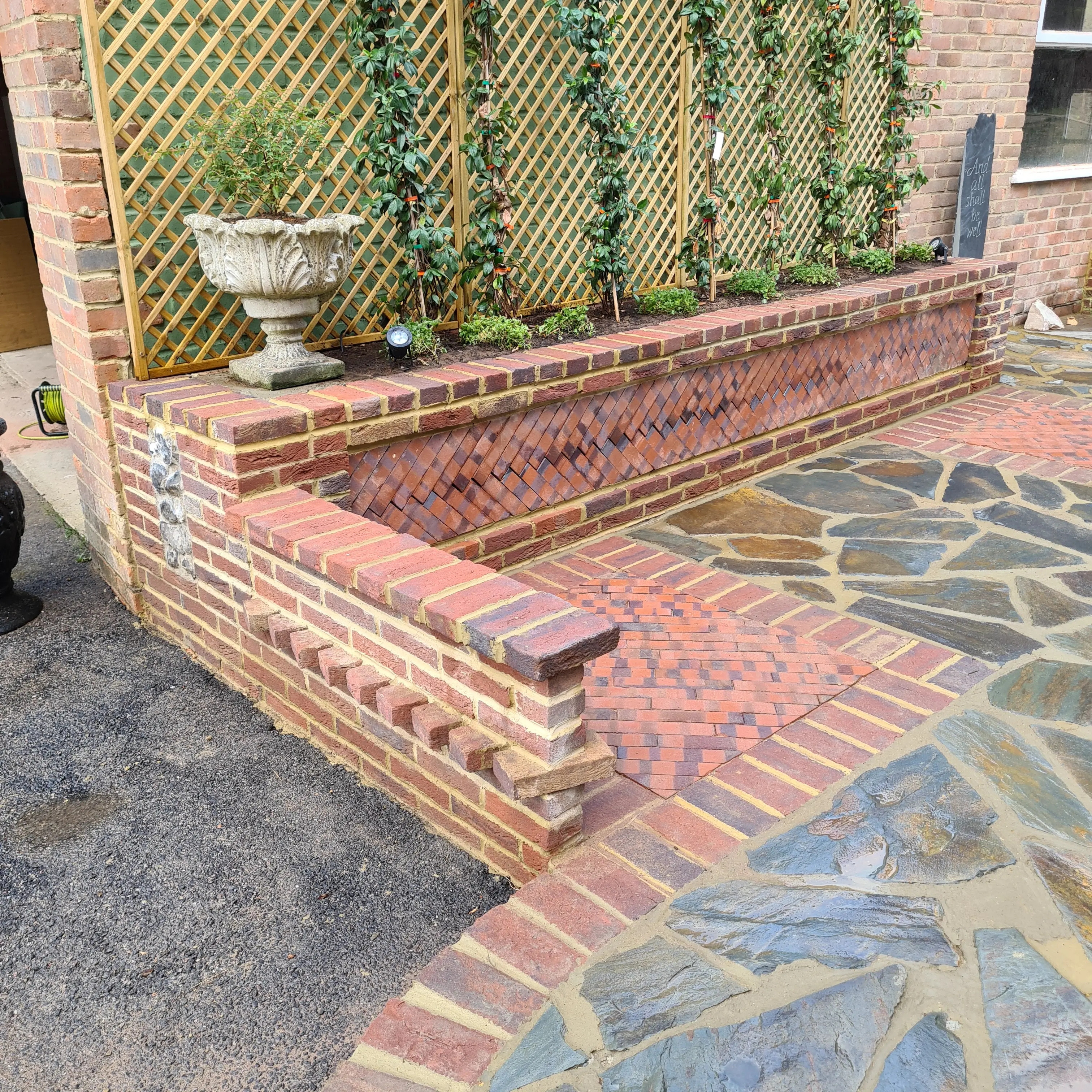 A brick seating area with a stone floor, surrounded by greenery, in a courtyard setting.