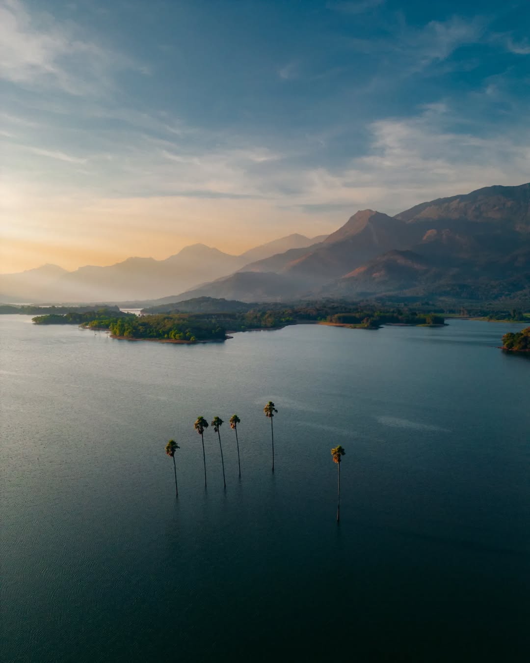 A breathtaking aerial view of Kava Island, surrounded by a vast expanse of water with a few palm trees emerging from the surface. The serene landscape is framed by distant mountains bathed in golden sunlight, creating a peaceful and picturesque scene