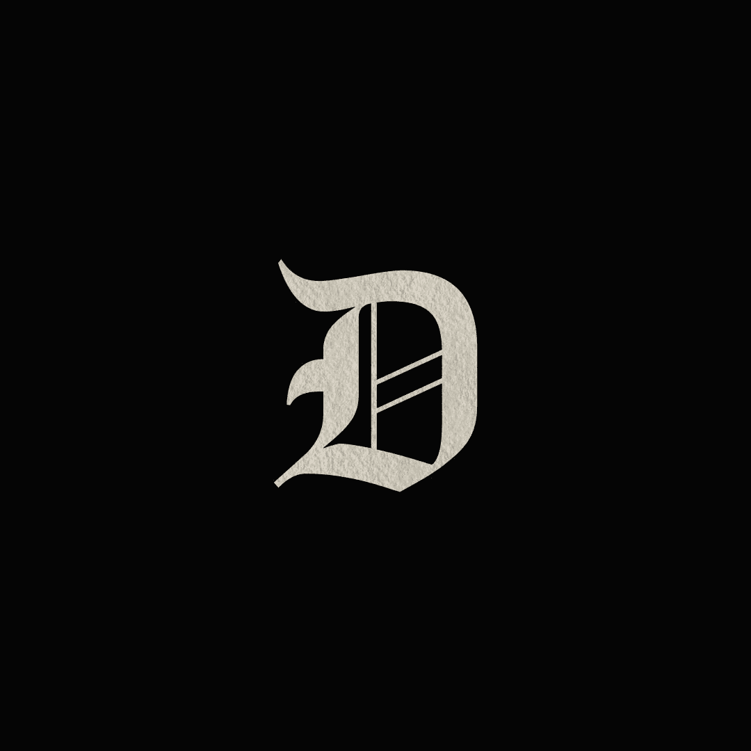 Woman in sunglasses poses against vibrant red background, wearing a beige blazer and black top. Sunlight casts dynamic shadows, creating a stylish, confident mood.