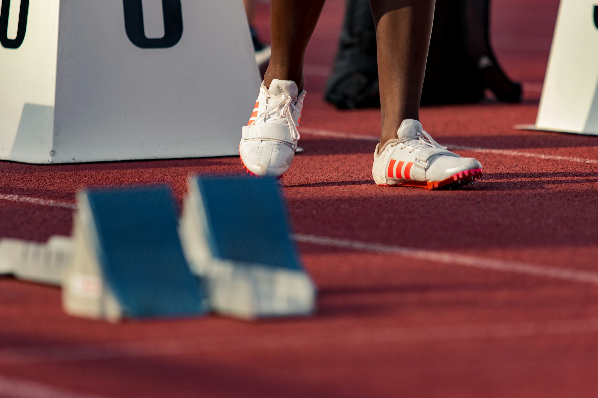 Close-up of an athlete's feet wearing white track spikes, standing next to blue starting blocks on a red track.
