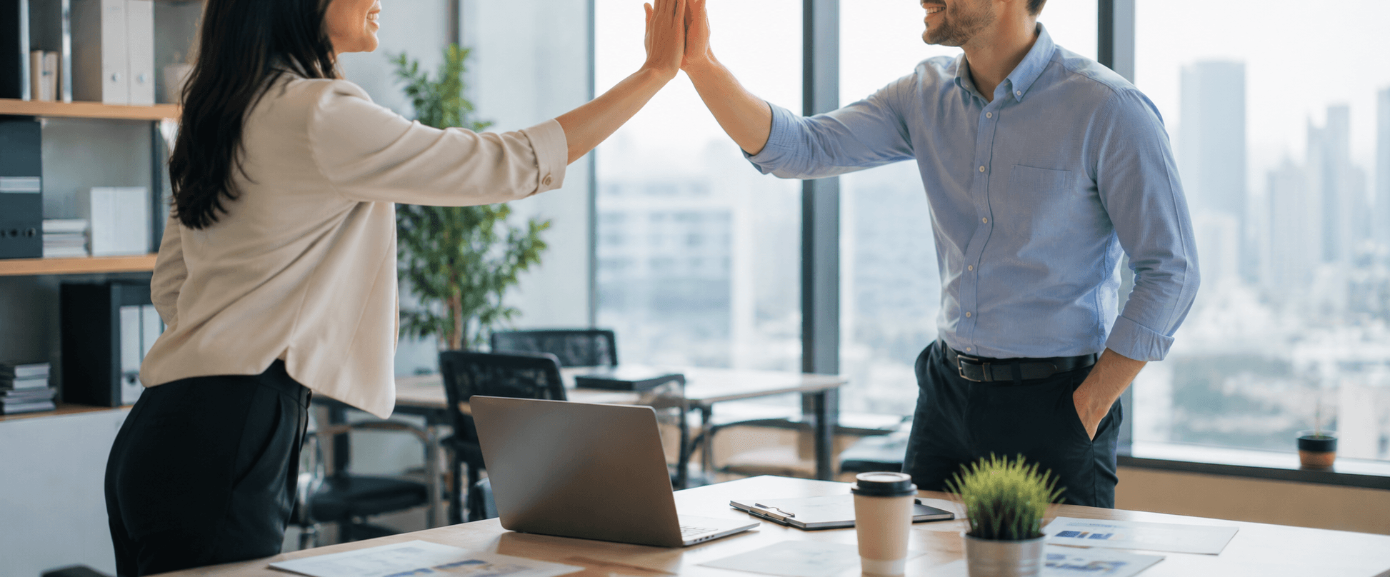 Two colleagues smiling and giving a high-five at a desk in a modern office with a laptop, coffee mug, and plants.