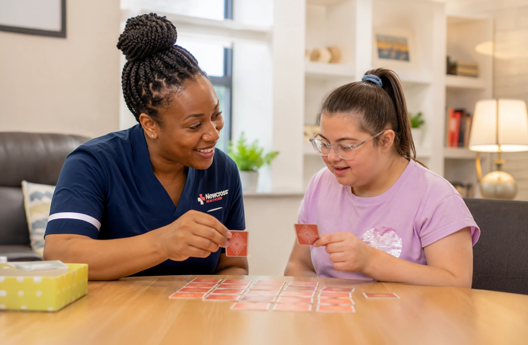 Nurse smiling and playing a card game with a young person with a disability in a warm, supportive care setting.
