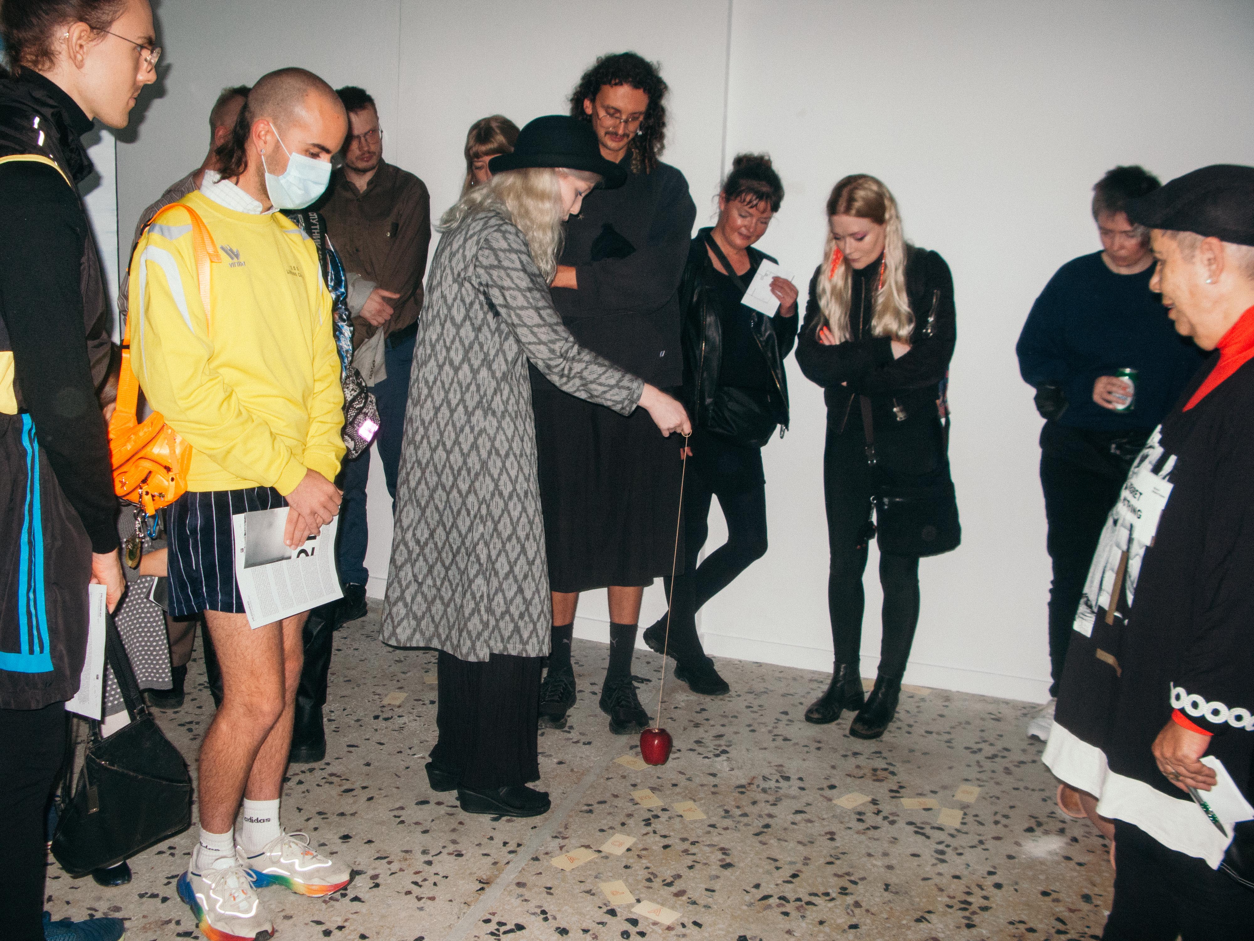 A still from Vowel step by Ásta Fanney Sigurðardóttir: the artist, in a patterned coat and dark hat, dangles a small red object on a string above lettered cards scattered across the floor, surrounded by a circle of attentive viewers.