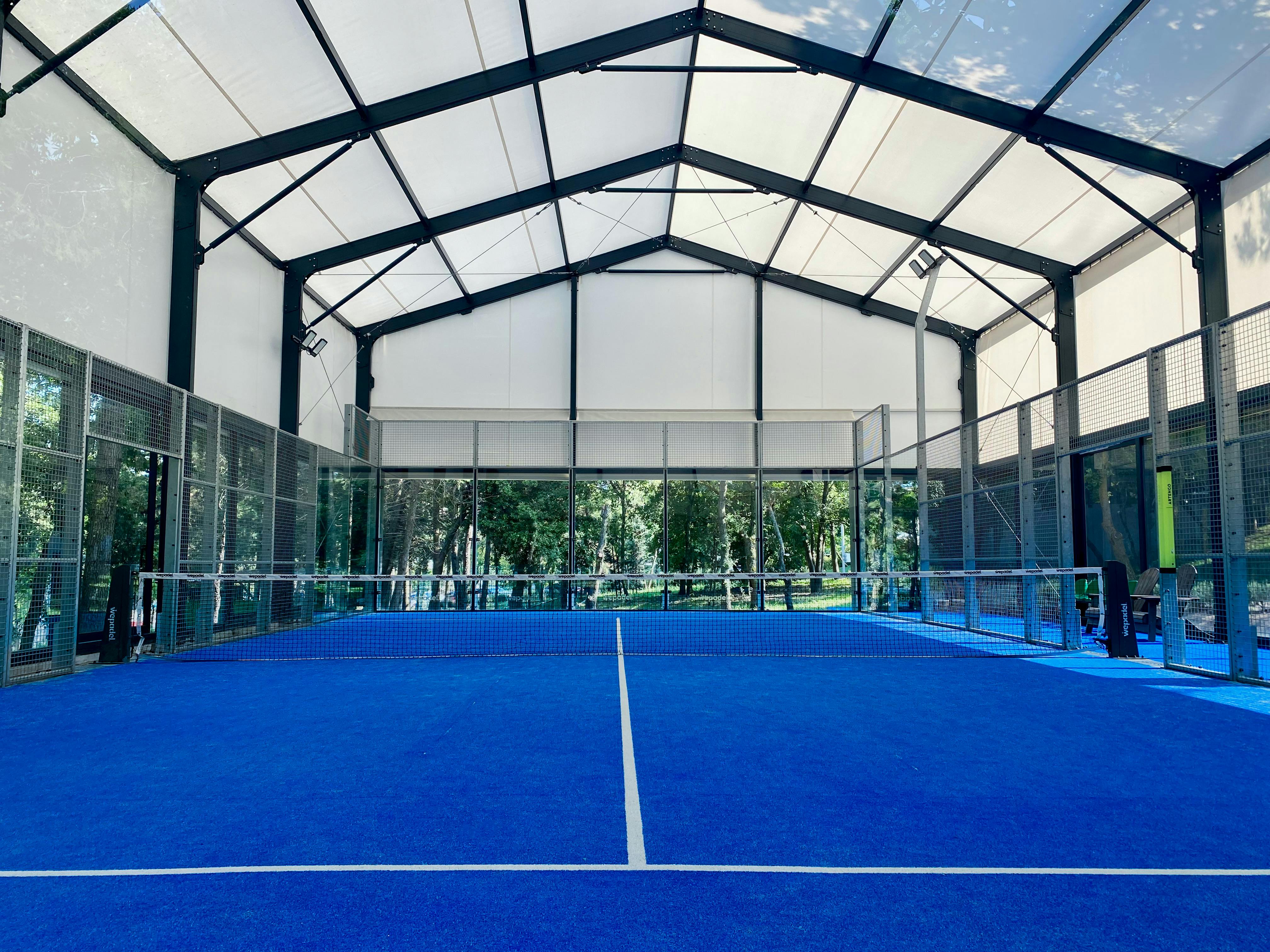 A modern, open-air padel court with a bright blue playing surface is enclosed by high, transparent walls and sheltered by a white canopy, surrounded by lush green trees.