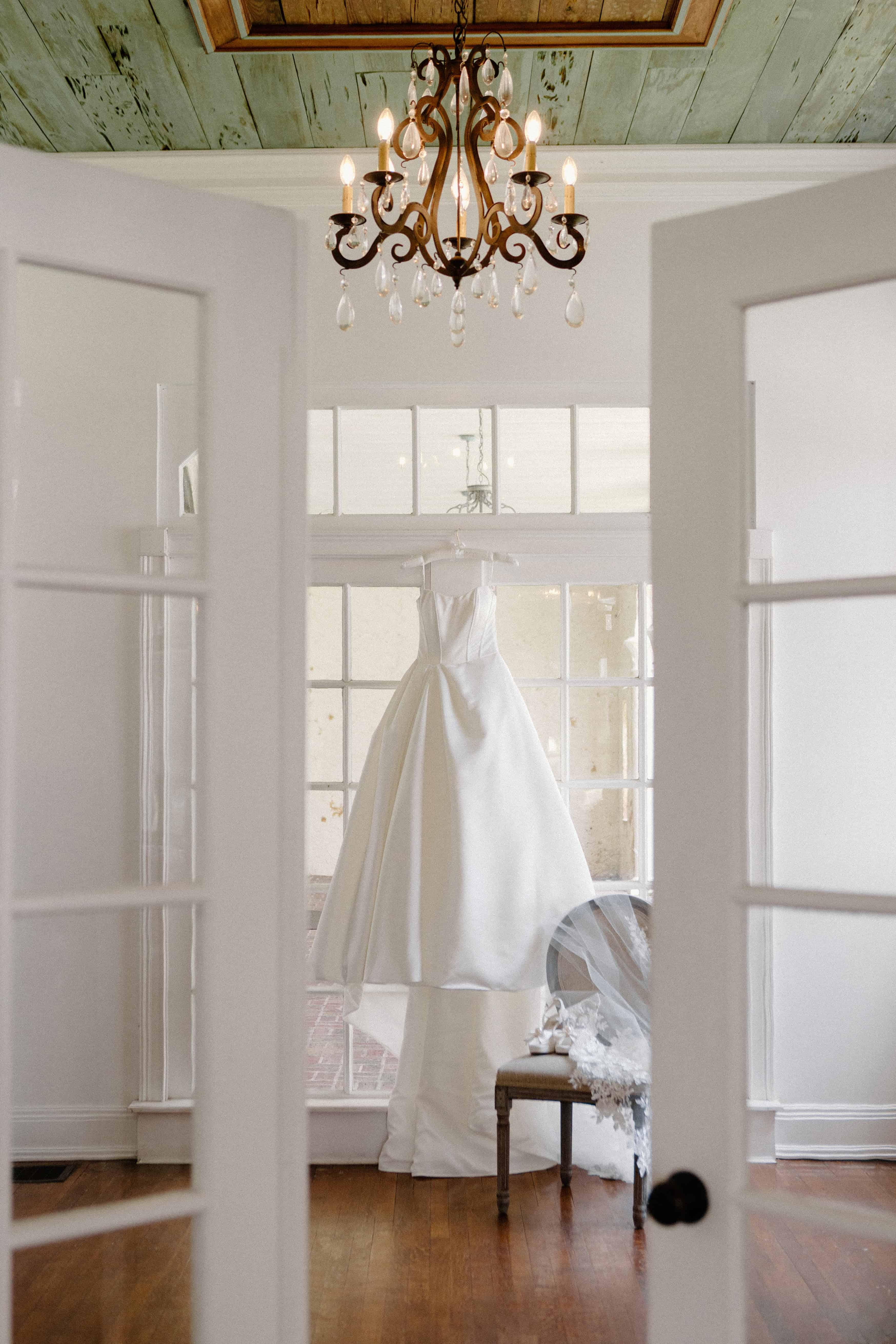 A photo of a white wedding dress hanging in a sunlit room, seen through an open doorway. A chandelier hangs from the ceiling.