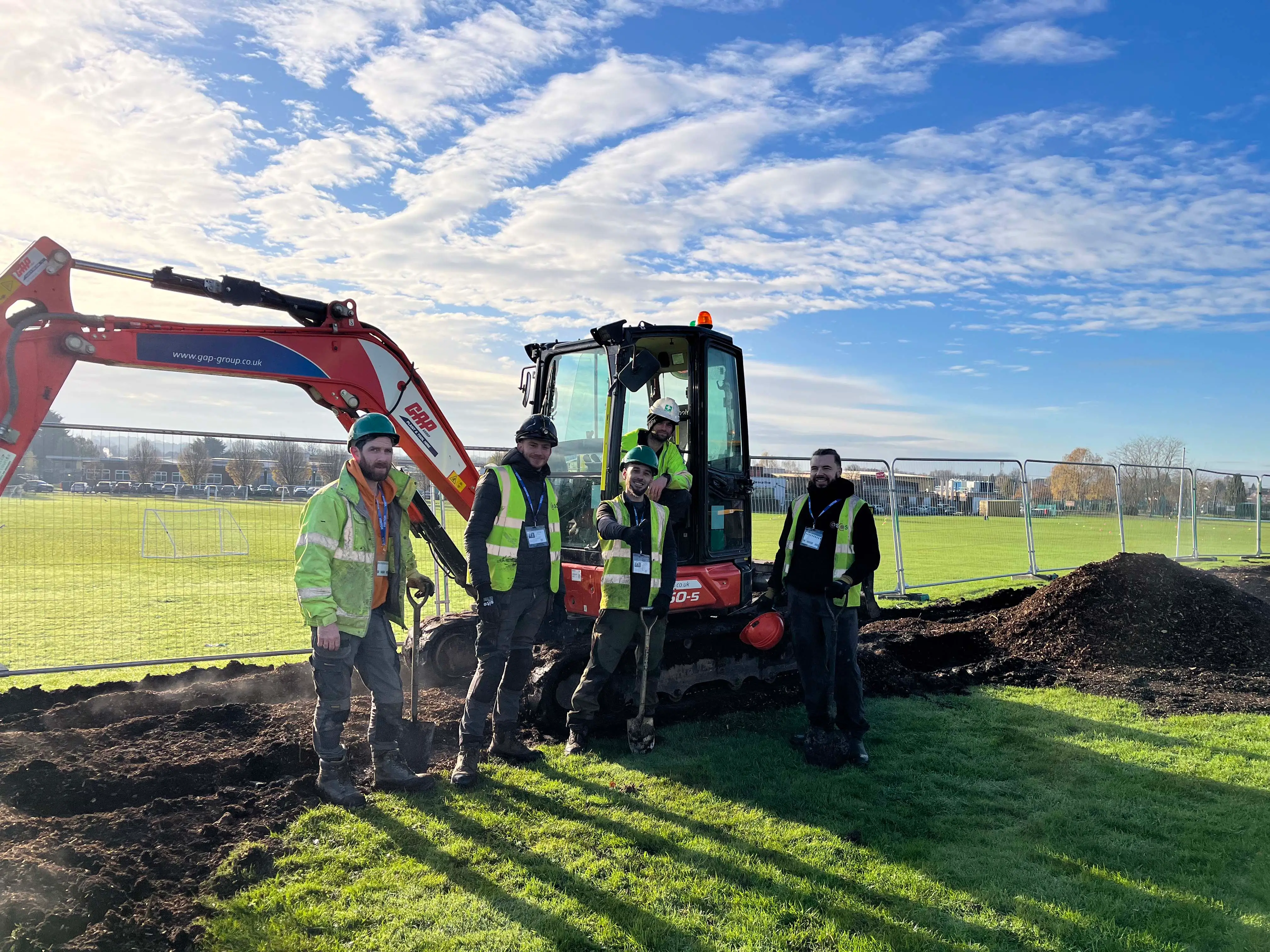 A tractor with an excavator attachment operates on a sunny field, with workers nearby. Blue sky with clouds above.