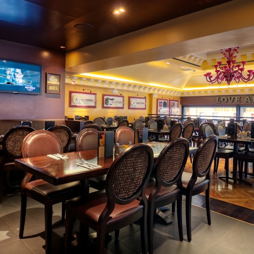 一家光线明亮的餐厅，配有木质餐桌、棕色椅子、壁挂电视、装裱画作和一盏粉色吊灯。