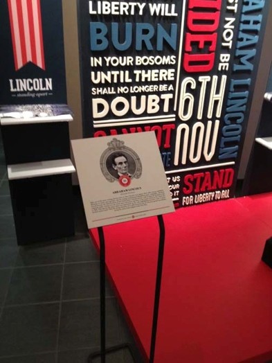 A stand depicting president Abe L. with some text underneath. Behind the stand there are r, w and b colored words