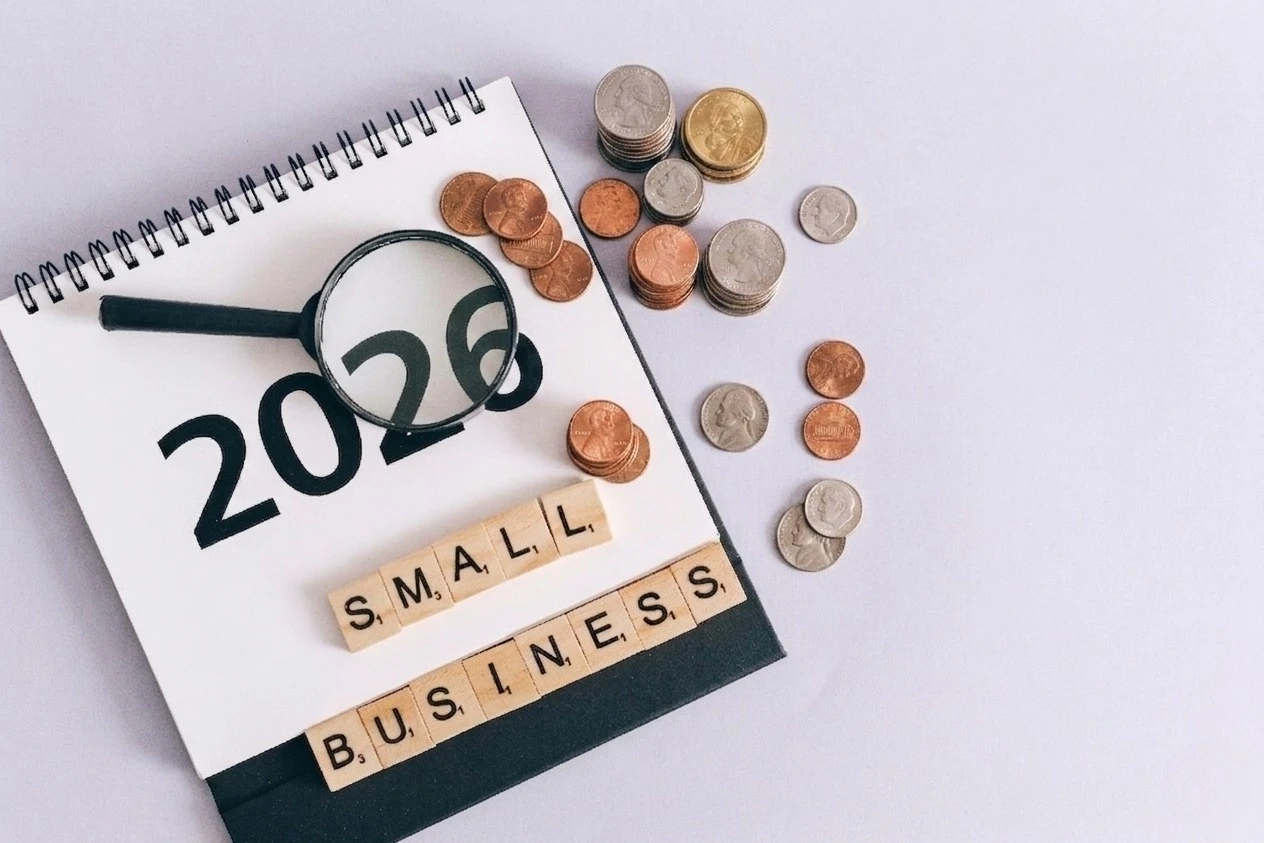 A magnifying glass resting on the year 2026 on a notepad, alongside wooden letter tiles spelling "SMALL BUSINESS" and scattered coins.