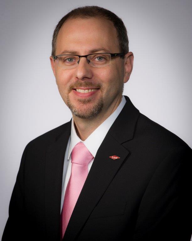 A smiling man wearing glasses and a formal suit with a pink tie poses in front of a neutral background.